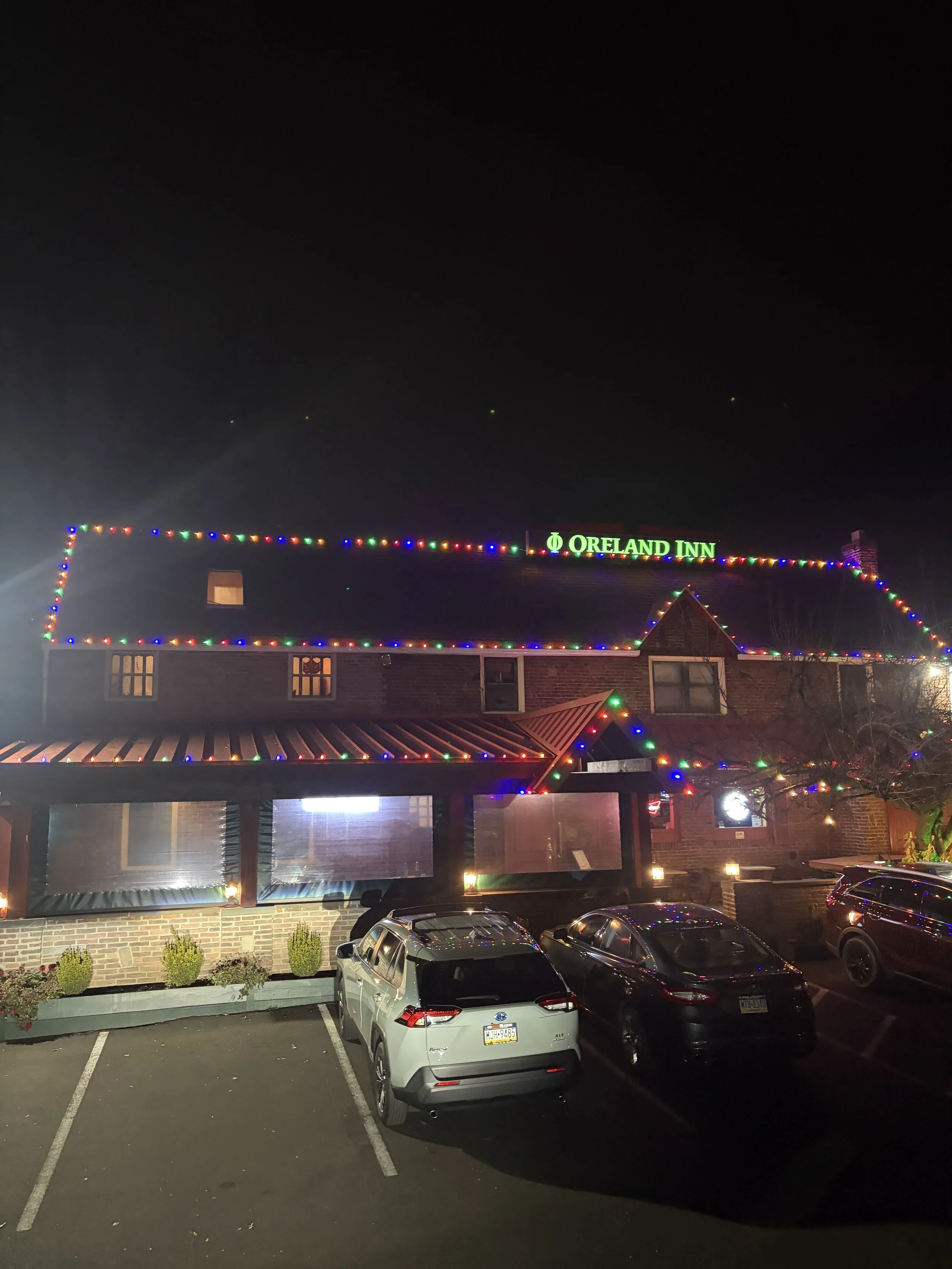 Night view of a building decorated with colorful Christmas lights and a green sign reading 'Oreland Inn', with a parking lot in front containing four cars.