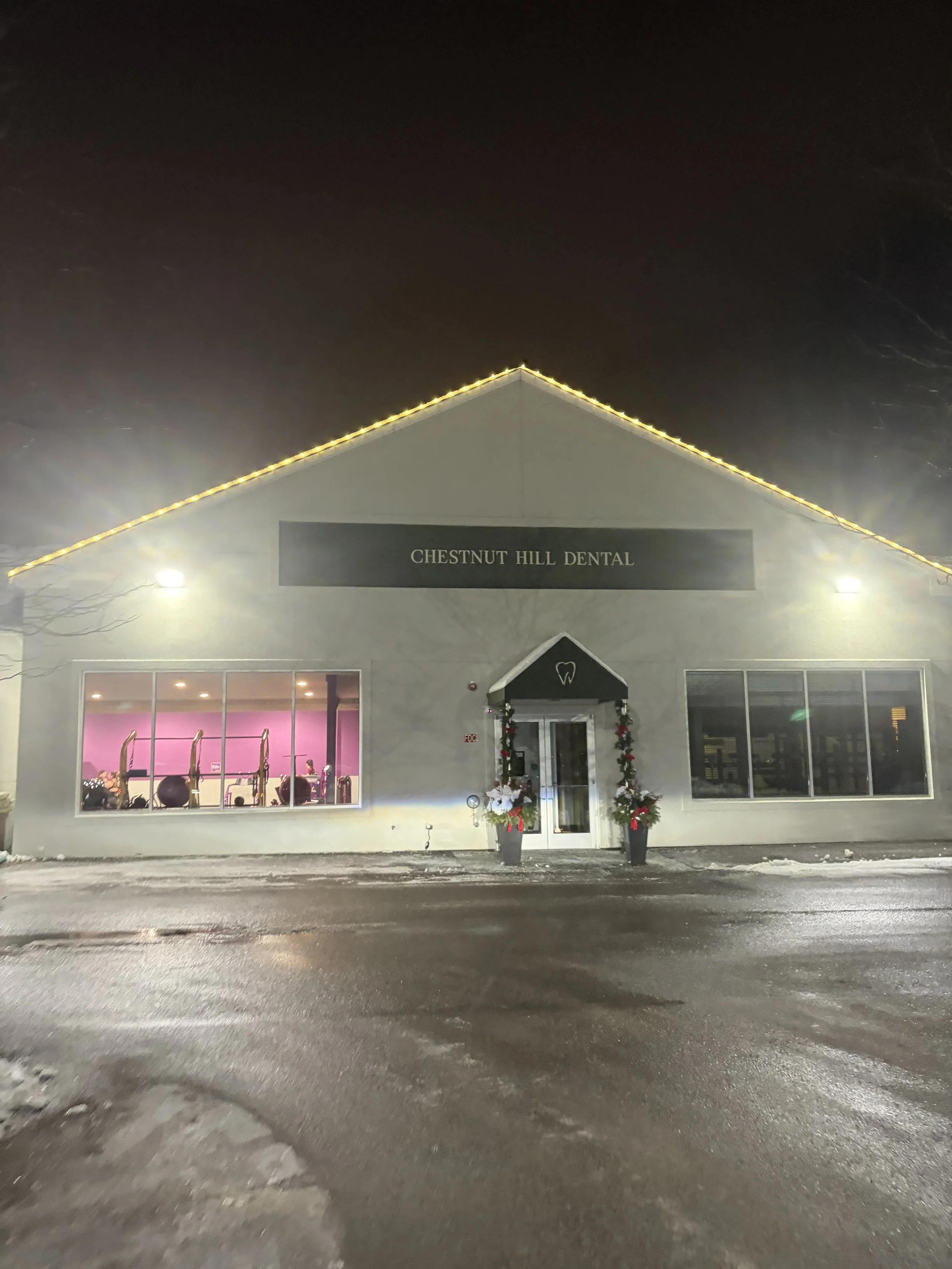Night view of a dental office named Chestnut Hill Dental with white exterior walls, decorated with Christmas garlands at the entrance, windows revealing a pink interior, and illuminated with yellow lights along the roofline. The parking lot is wet, reflecting lights, and some snow is present on the ground.