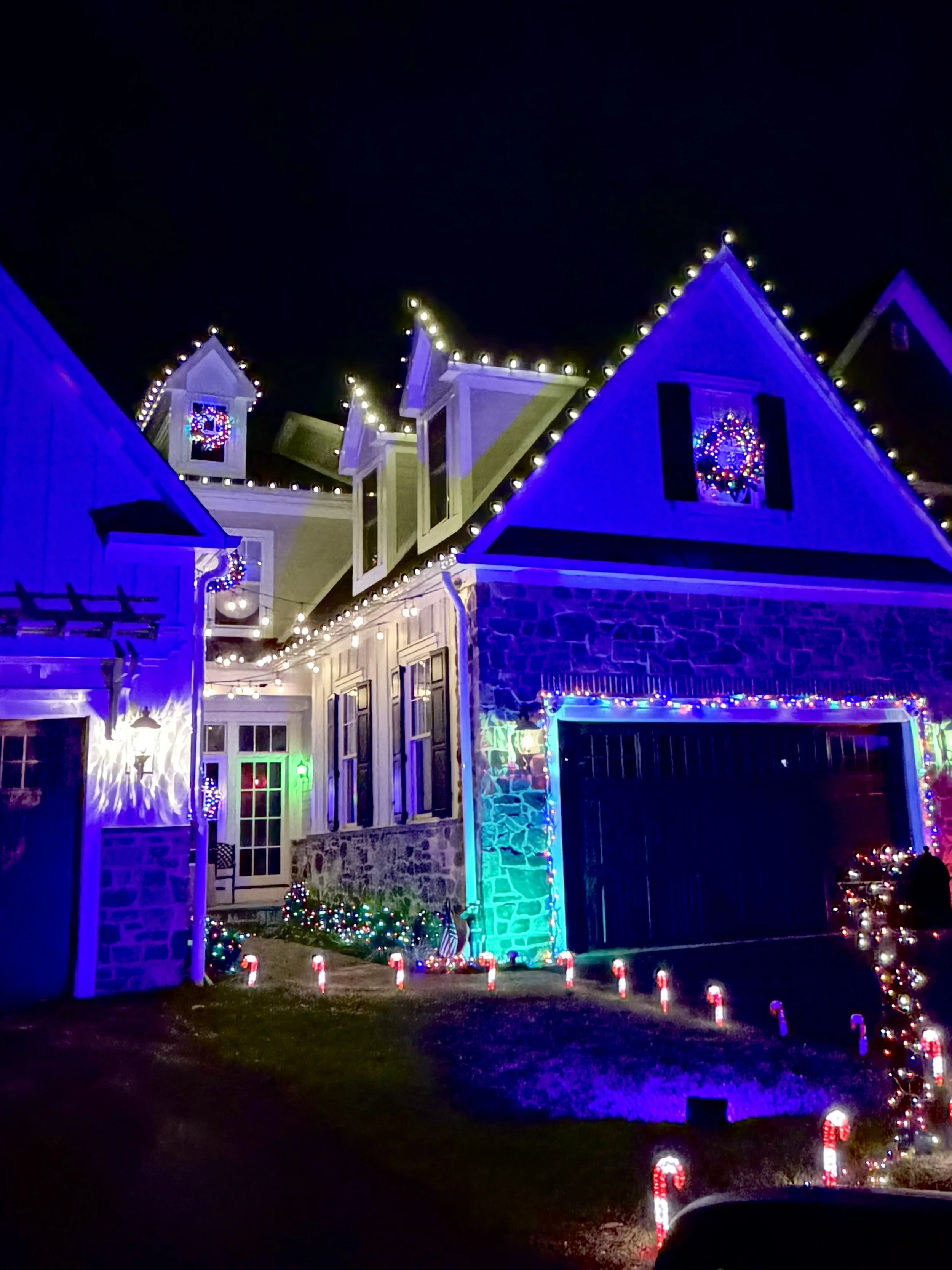 A house decorated for Christmas with multicolored lights outlining the roof, windows, and entryway, and a wreath on the window. Candy cane solar lights line the driveway, and Christmas trees with lights are visible, all against a dark night sky.