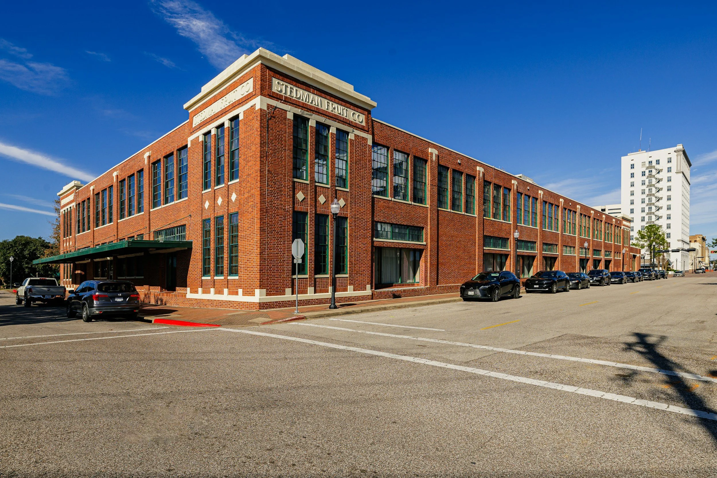 A large red brick building with multiple windows and a sign that reads "STEDMAN FRUIT CO" on the corner. The street in front has parked cars and a clear blue sky overhead.