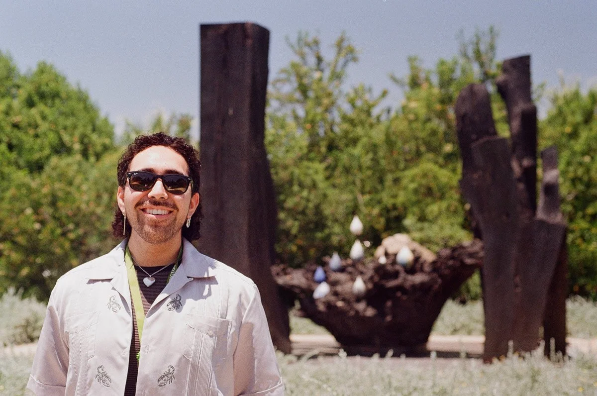 A man smiling wearing sunglasses, earrings, and a white embroidered shirt, standing outdoors in front of an art installation with tree-like structures and ceramic objects.