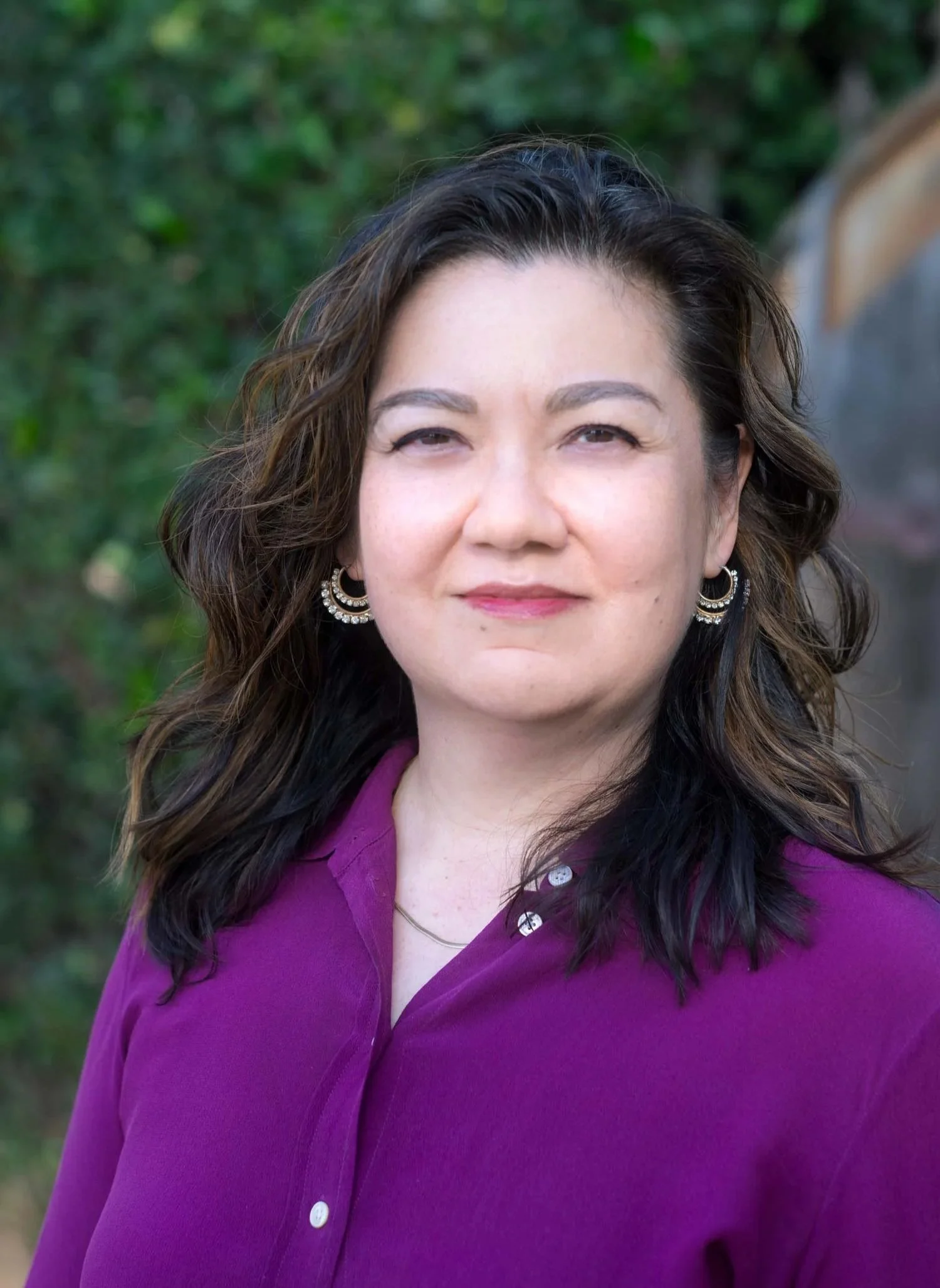 Close-up of a woman with dark wavy hair, wearing a purple blouse and large hoop earrings, outdoors with green foliage in the background.