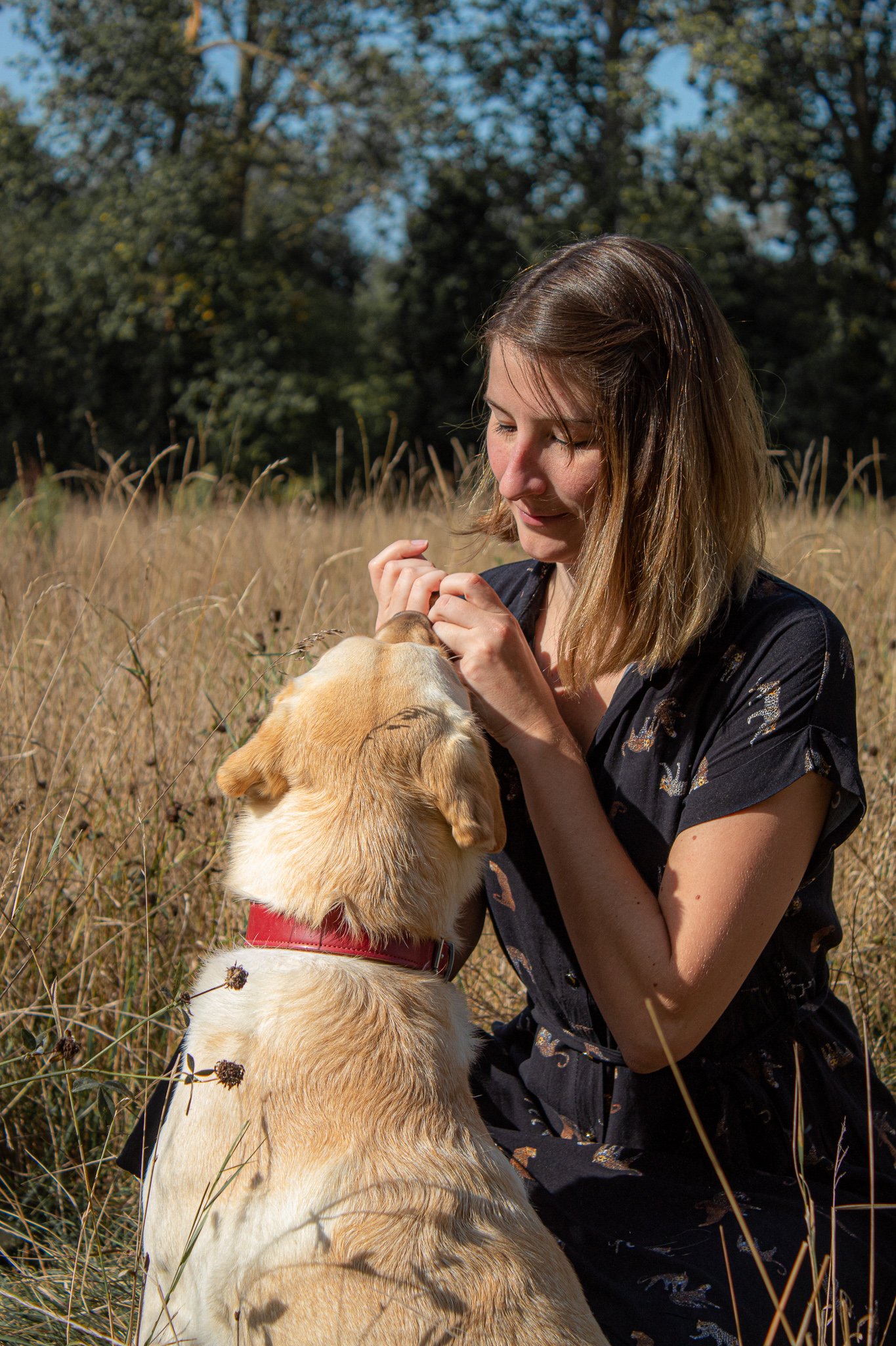 Een jonge vrouw met bruin haar knielt in een veld met lang gras, en interacteert met een golden retriever hond.