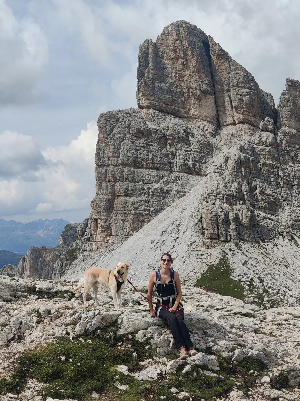 Vrouw en hond op een berg met grote rotsformatie op de achtergrond, bewolkte hemel.