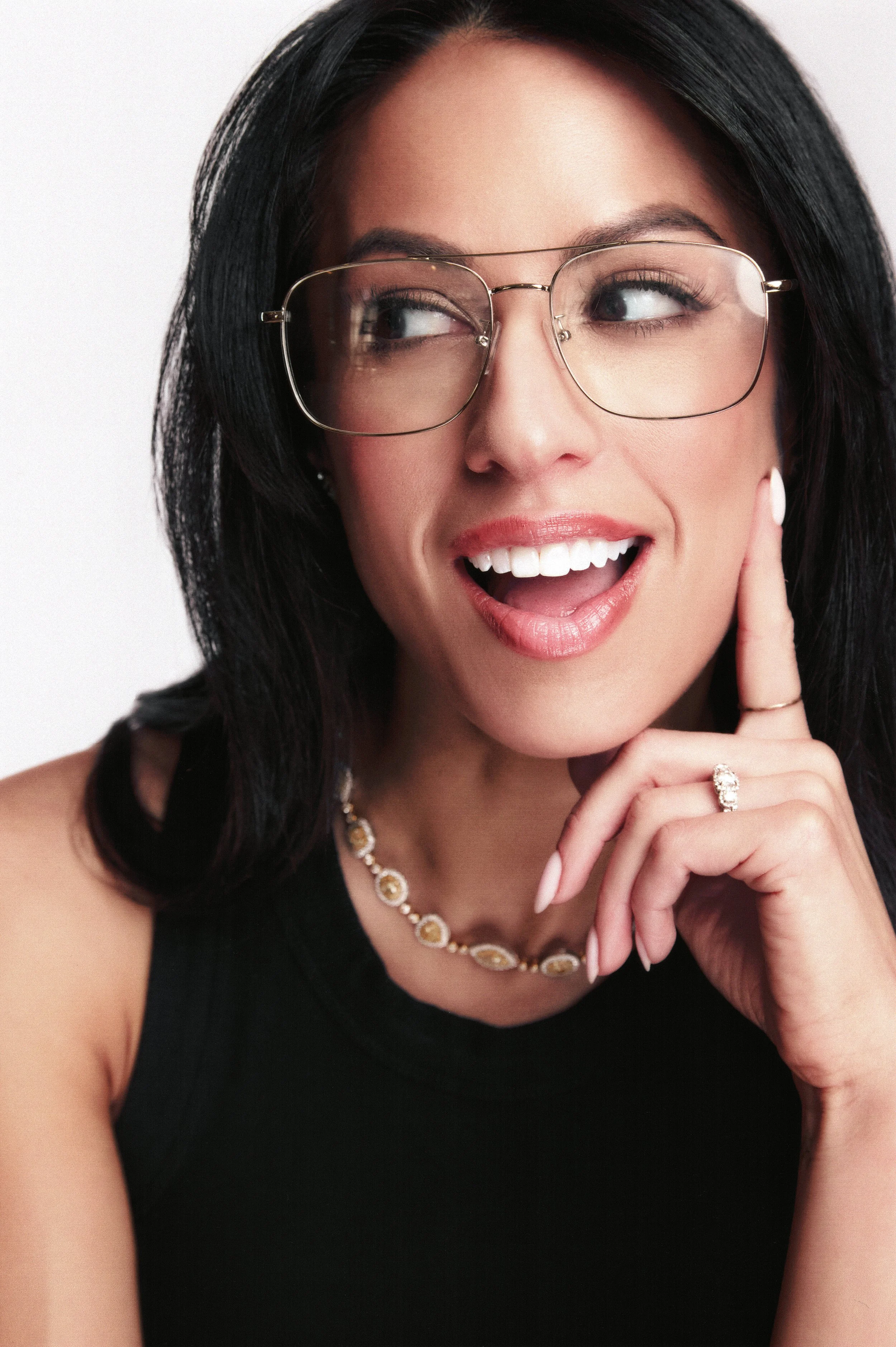 A woman with long black hair wearing glasses, a black top, and jewelry, smiling with her hand touching her face.