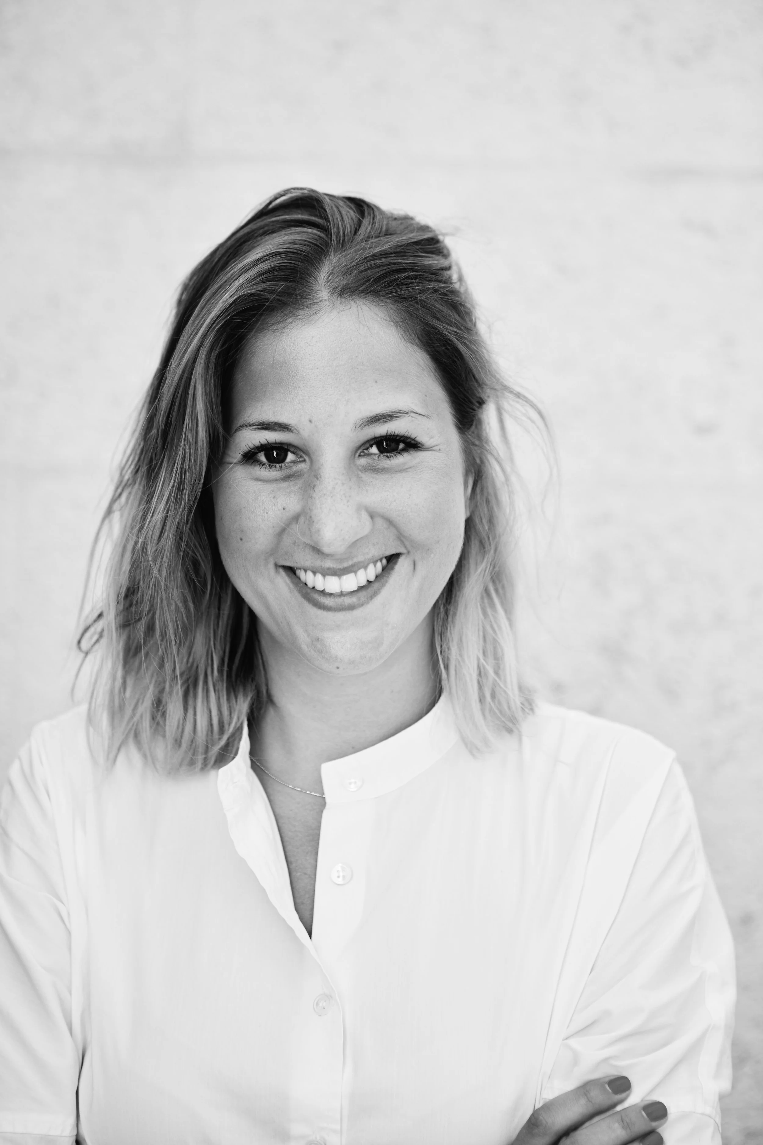 Schwarz-Weiß-Foto einer lachenden jungen Frau mit mittelgroßen Haaren, die eine weiße Bluse trägt, vor hellem Hintergrund.