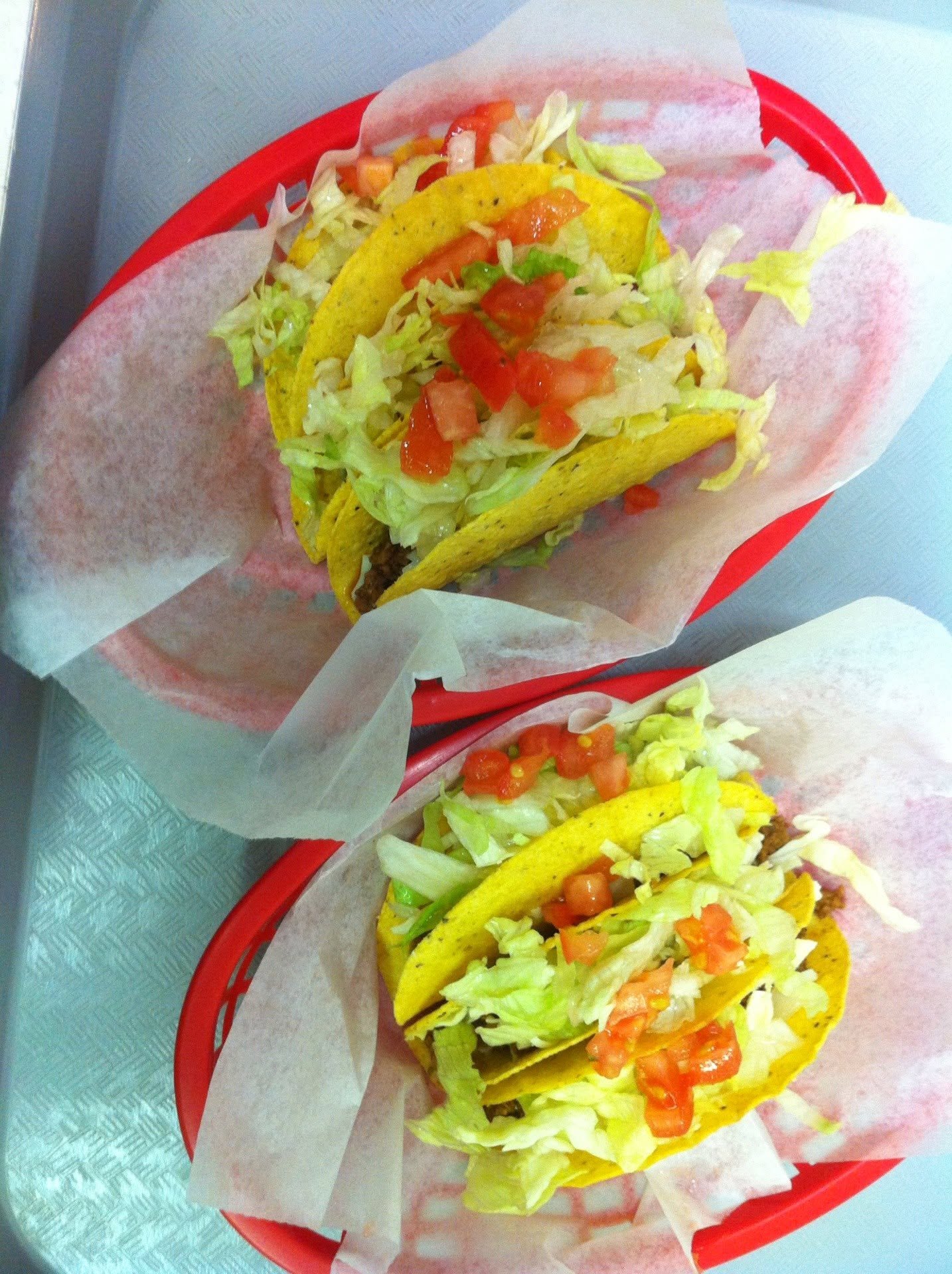 Two tacos with lettuce, diced tomatoes, shredded cheese, and taco meat on yellow corn tortillas, served in red baskets with paper lining.