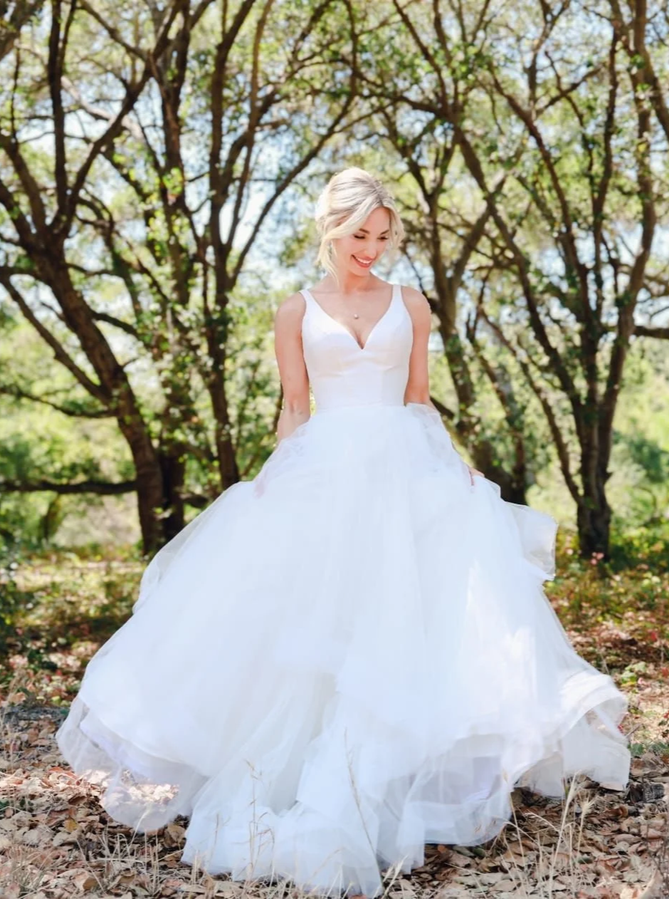 A bride in a white wedding gown standing outdoors among trees with green leaves, smiling and looking down.