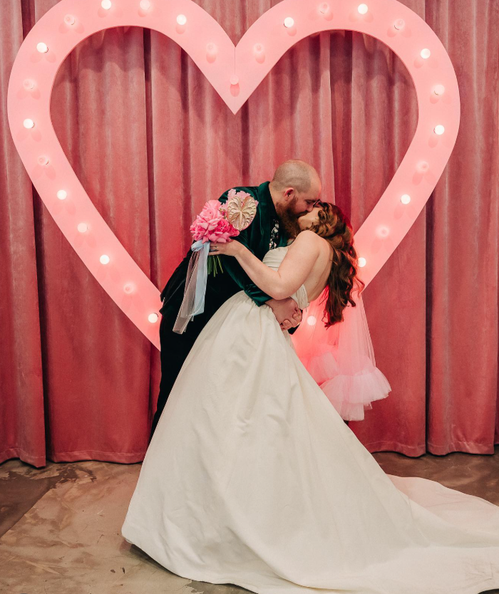 A bride and groom share a kiss in front of a large, illuminated pink heart on a pink curtain backdrop at their wedding.