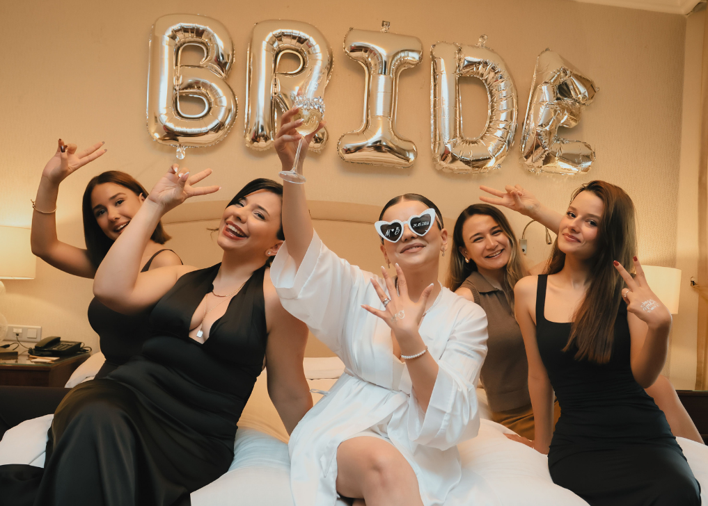 Five women celebrating a birthday on a bed, with silver balloon letters spelling 'BIRTHDAY' in the background.