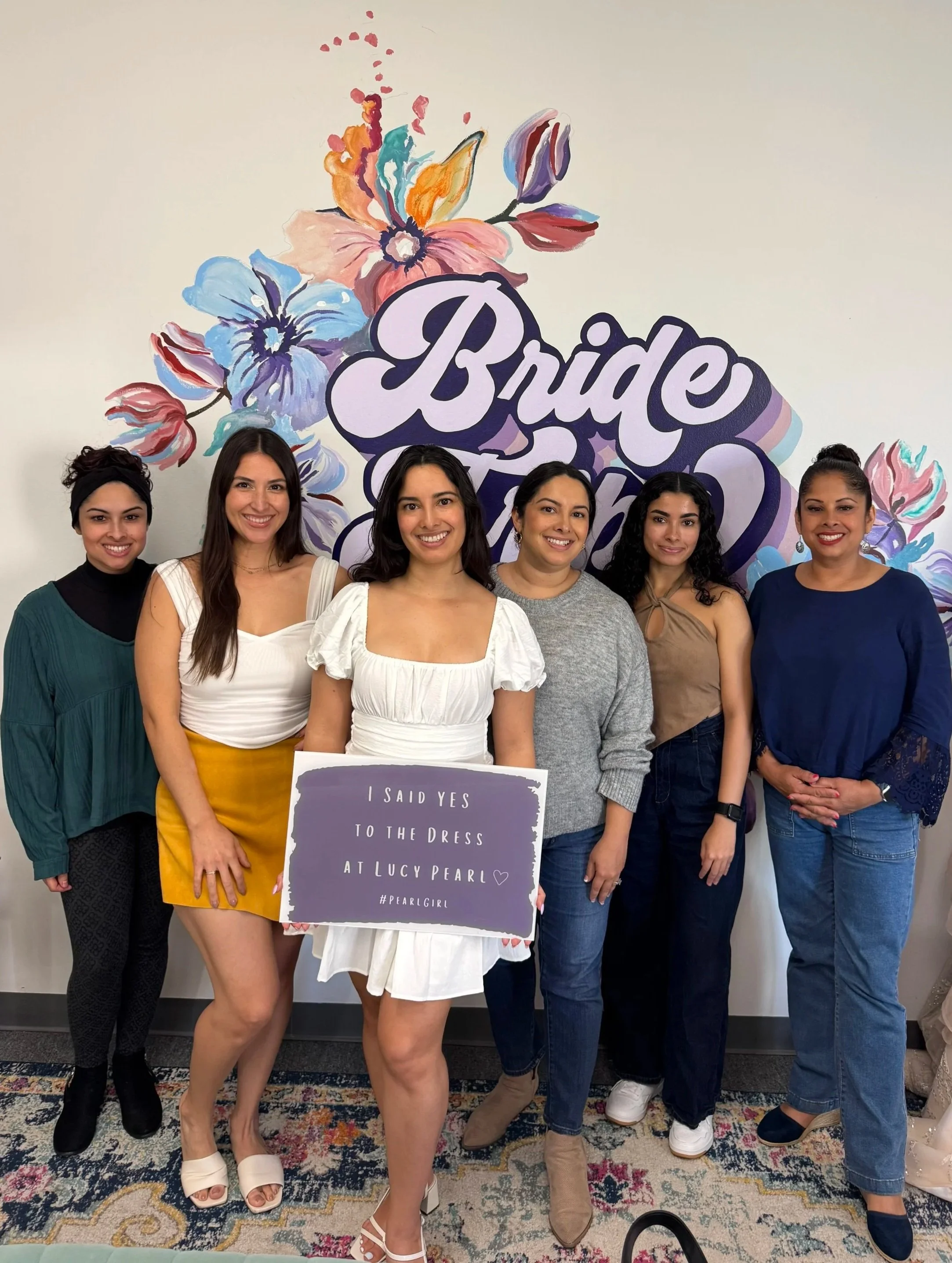 Group of six women standing together in front of a colorful floral background with the words "Bride Tribe" visible. One woman holds a sign that reads, "I said yes to the dress at Lucy Pearl."