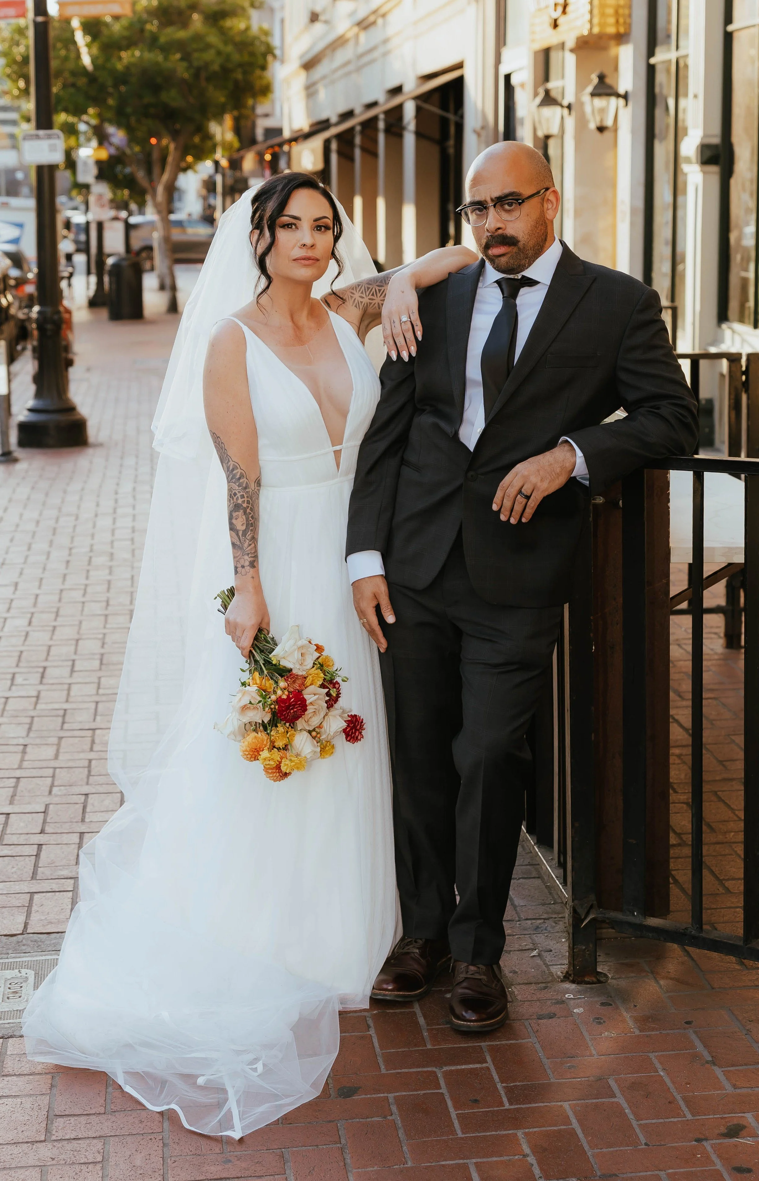 A bride and groom posing together outdoors on a city street. The bride is wearing a white wedding dress and holding a bouquet of flowers, with her arm resting on the groom's shoulder. The groom is dressed in a dark suit and glasses, leaning on a railing with a serious expression.