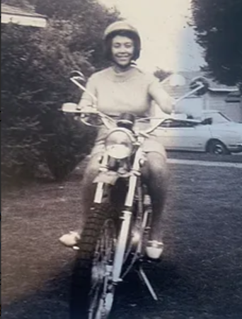 Black and white photo of a woman smiling while sitting on a bicycle outdoors, with cars and trees in the background.