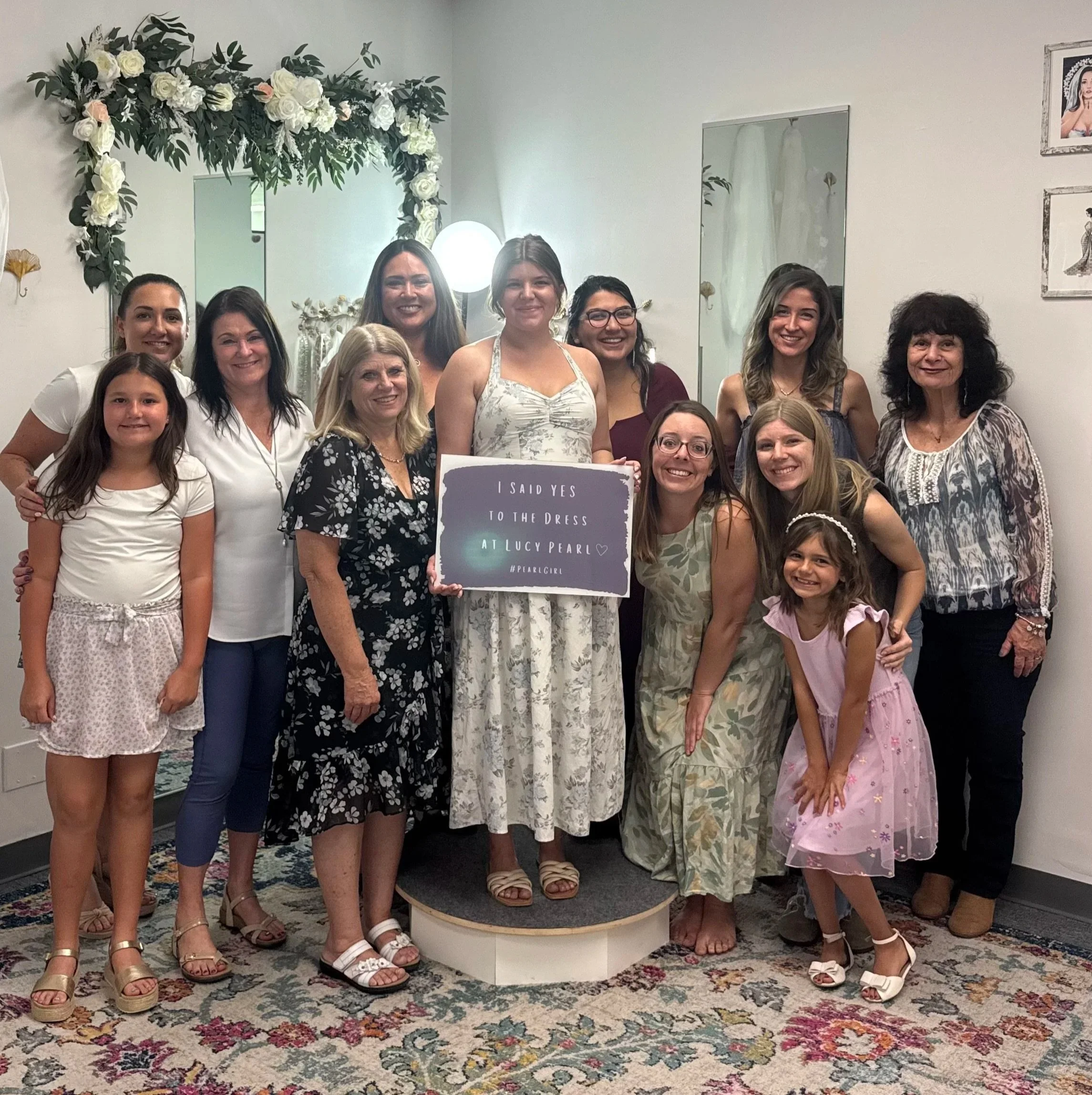 Group of women and girls posing together on a floral rug in front of a decorated wall with flowers and a mirror at a celebration or event. Some are smiling, and one woman holds a sign that says 'I said yes to the dress'.