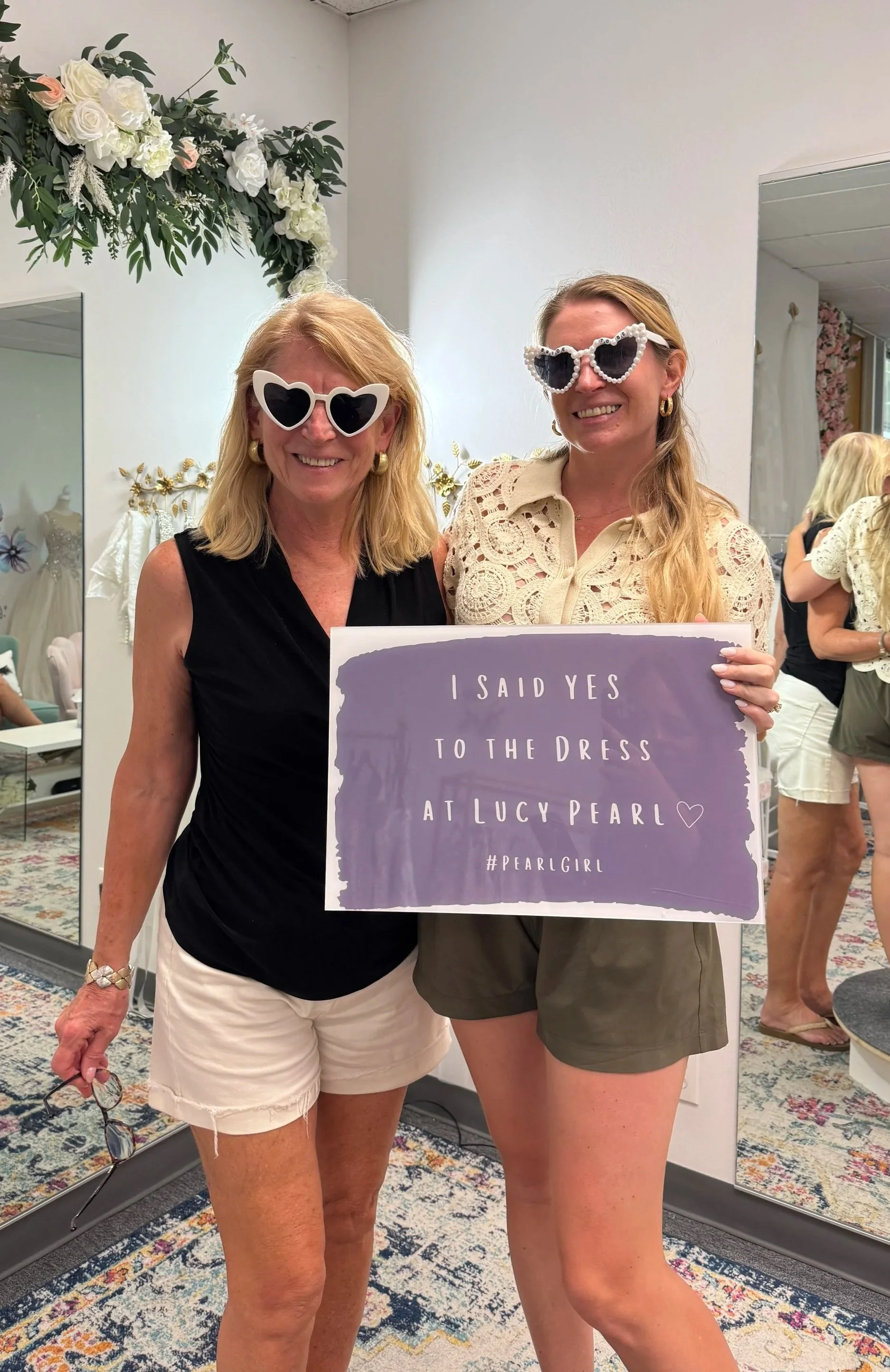 Two women wearing sunglasses while holding a sign that reads 'I said yes to the dress at Lucy Pearl,' standing in a floral decorated room, possibly a boutique.