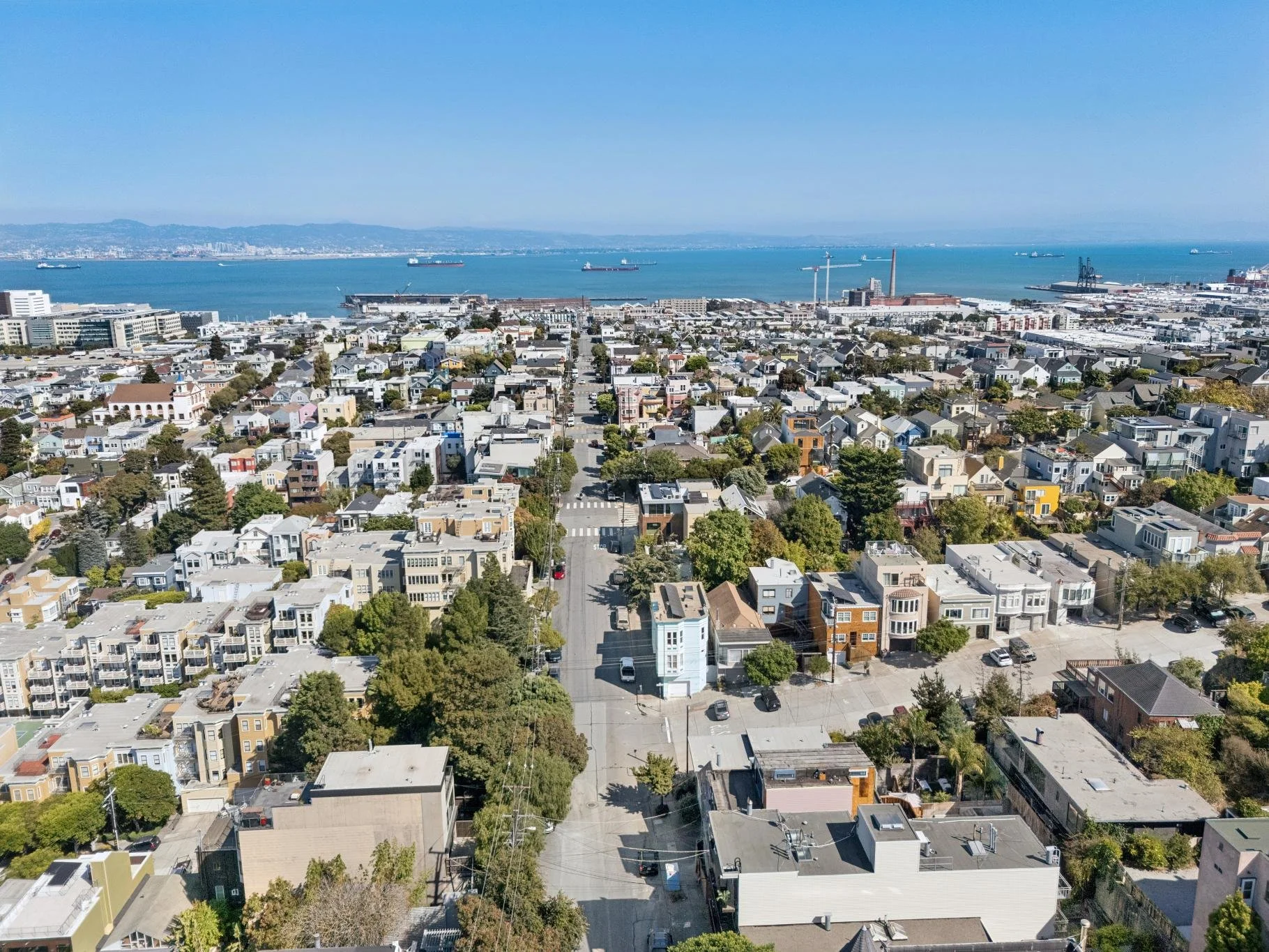 Aerial view of a coastal city with residential houses, commercial buildings, and street grid, overlooking the ocean with ships and a distant shoreline.