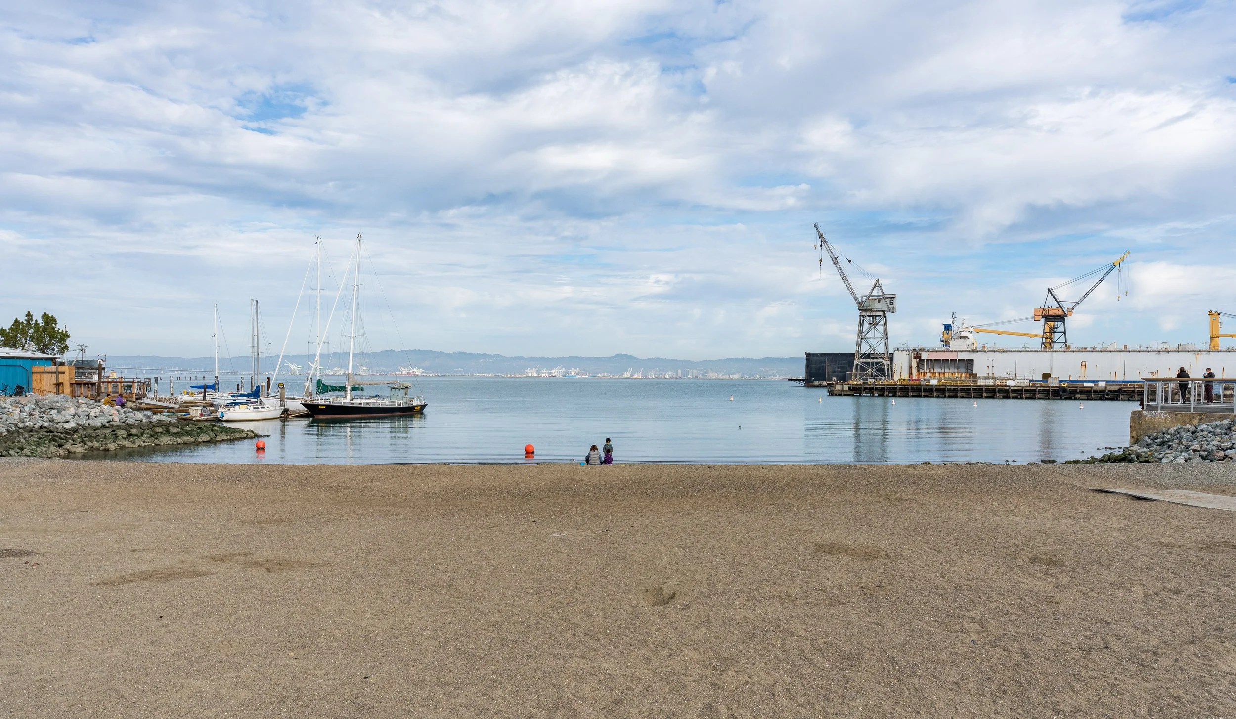 A waterfront scene with a sandy beach in the foreground, boats docked at a pier on the left, industrial cranes and a ship on the right, and a city skyline across the water under a partly cloudy sky.
