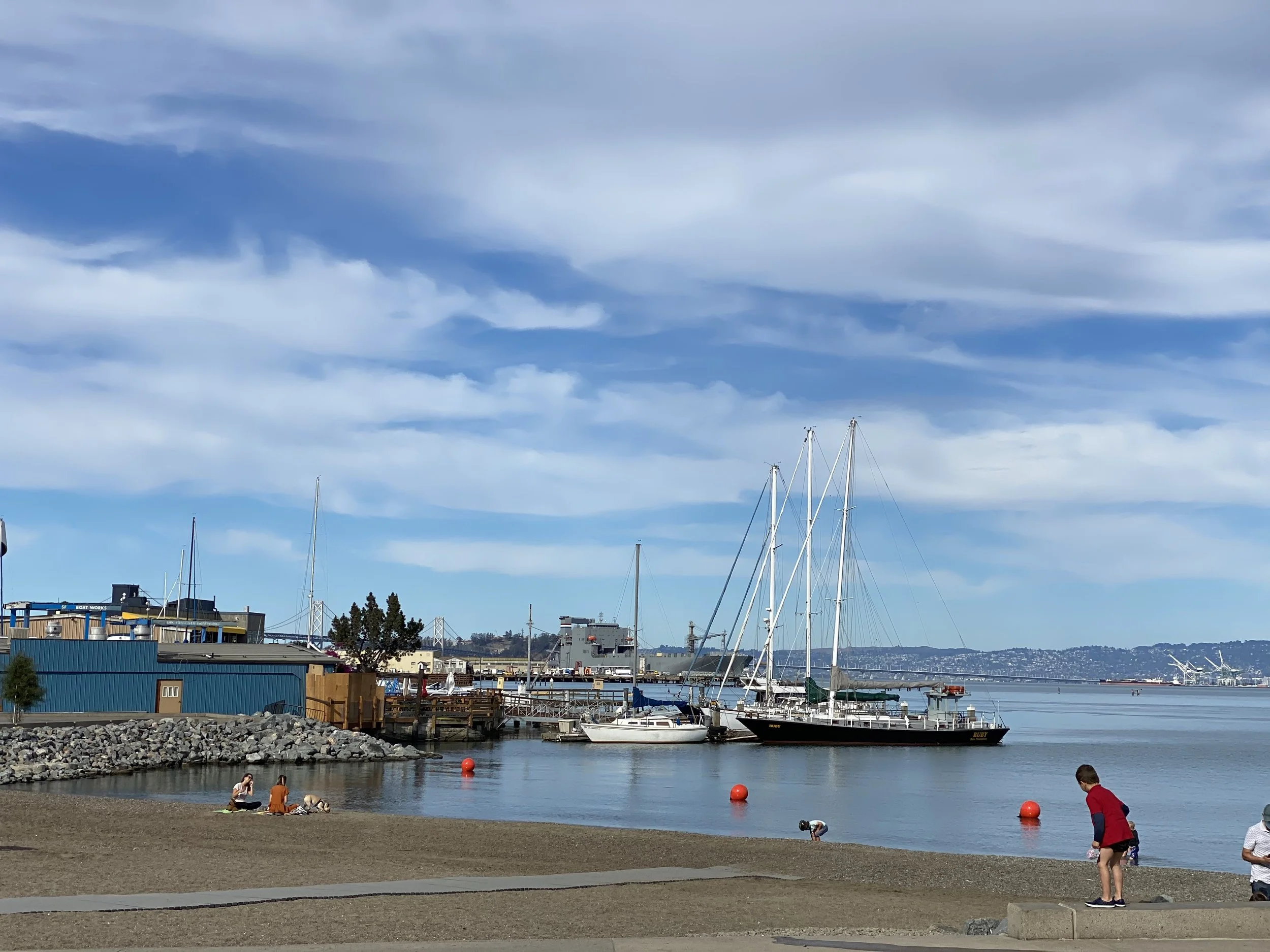 A marina with sailboats docked and people relaxing by the water, under a partly cloudy sky.
