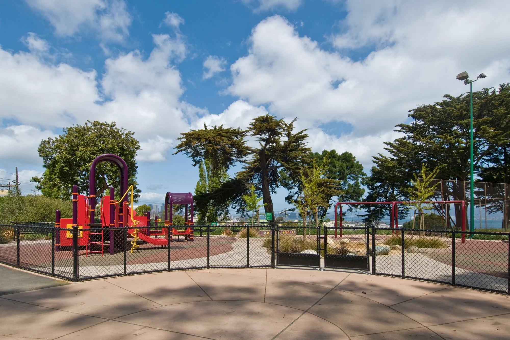 An empty outdoor playground with colorful play structures and equipment, surrounded by a black fence. Tall trees and a partly cloudy blue sky are in the background.