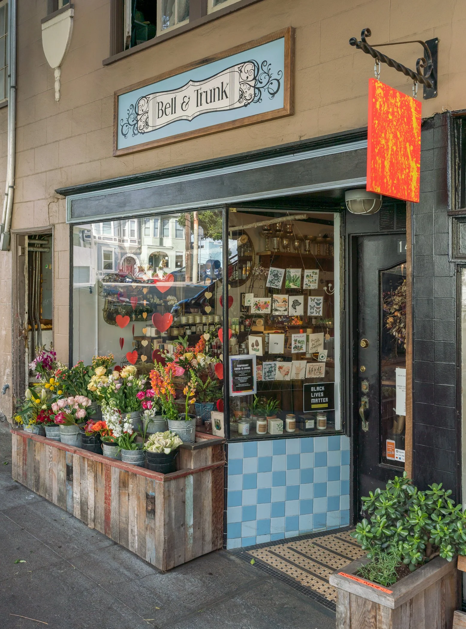 The storefront of a small flower and gift shop called "Bell & Trunk." It has a large window displaying flowers and greeting cards. Outside, there are potted flowers, a black door with a sign, and a small wooden planter box with green plants. A sign i