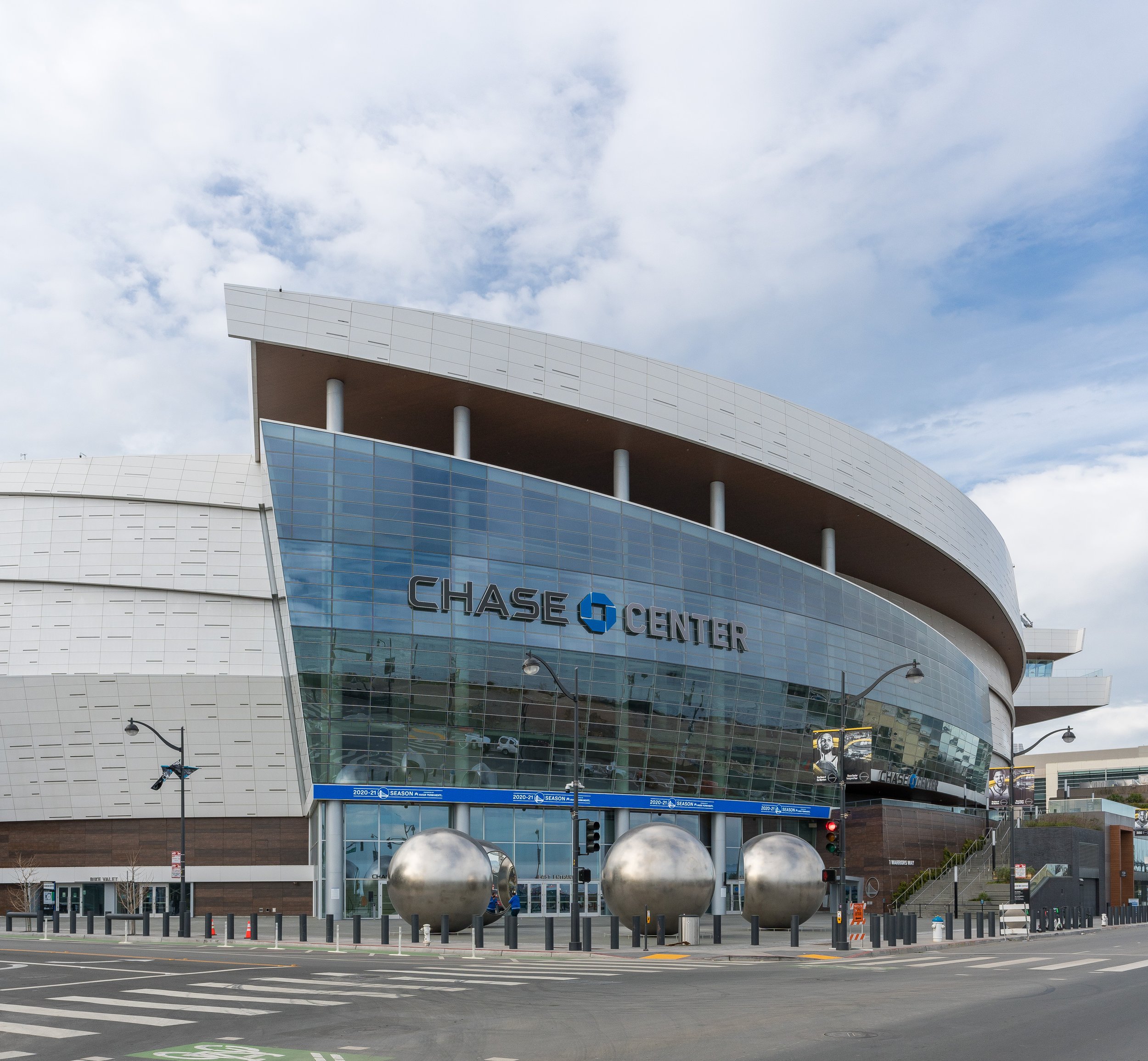 Exterior of the Chase Center arena with modern architectural design, glass windows, and metallic accents, under a cloudy sky.