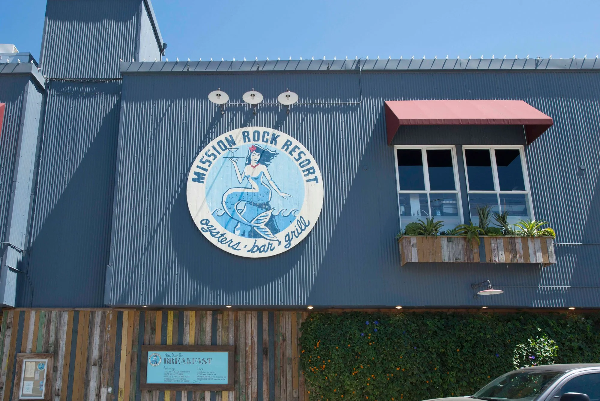 Exterior view of a building painted in dark blue with a large sign that reads 'Mission Rock Resort' featuring a mermaid holding a tray. The building has a red awning over a window, a wooden panel fence at the bottom, and some plants and flowers under