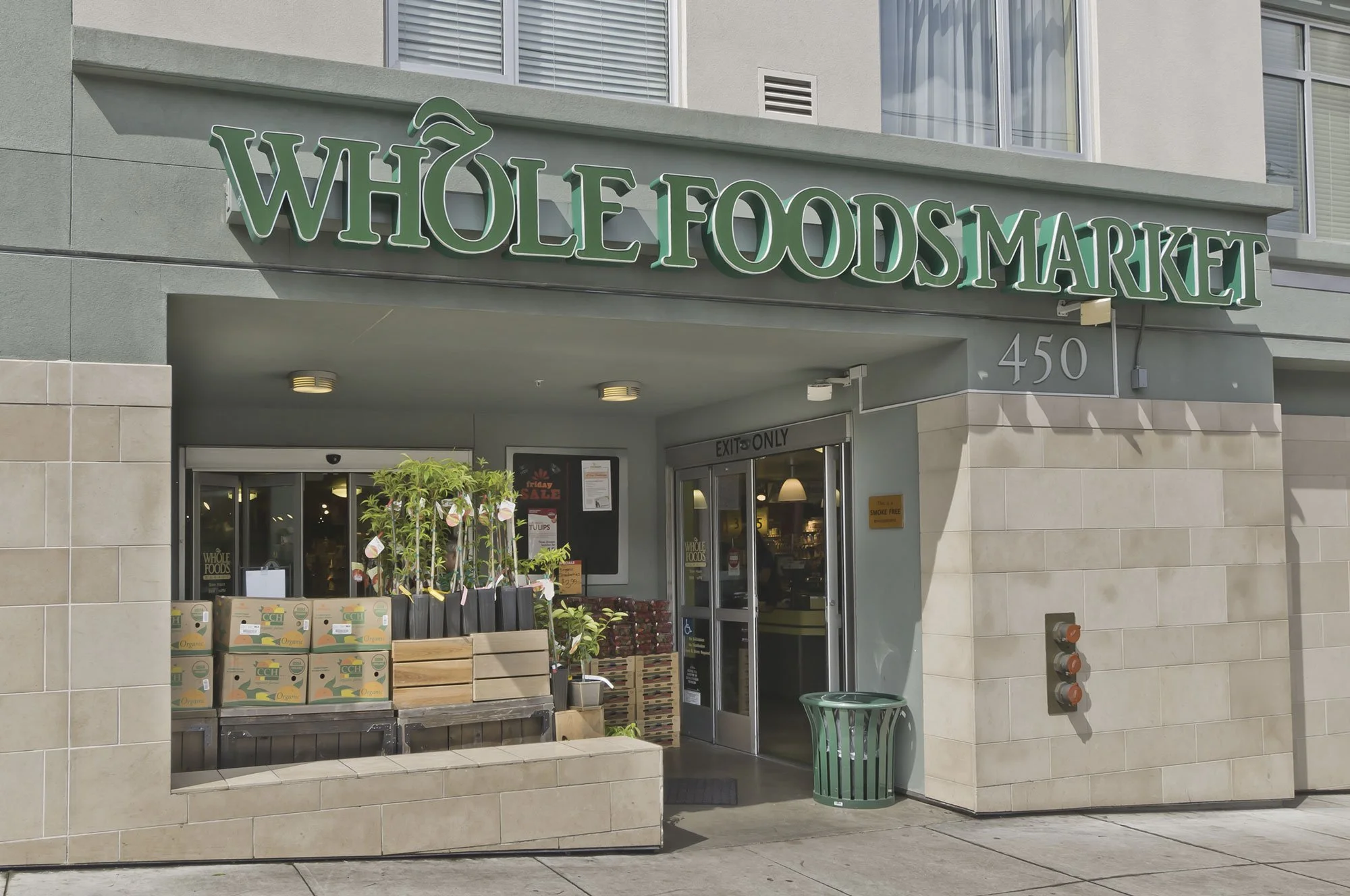 Entrance of Whole Foods Market with stacked boxes of organic produce, green plants, and a trash can outside. Sign with green letters displays 'Whole Foods Market' above the entrance.