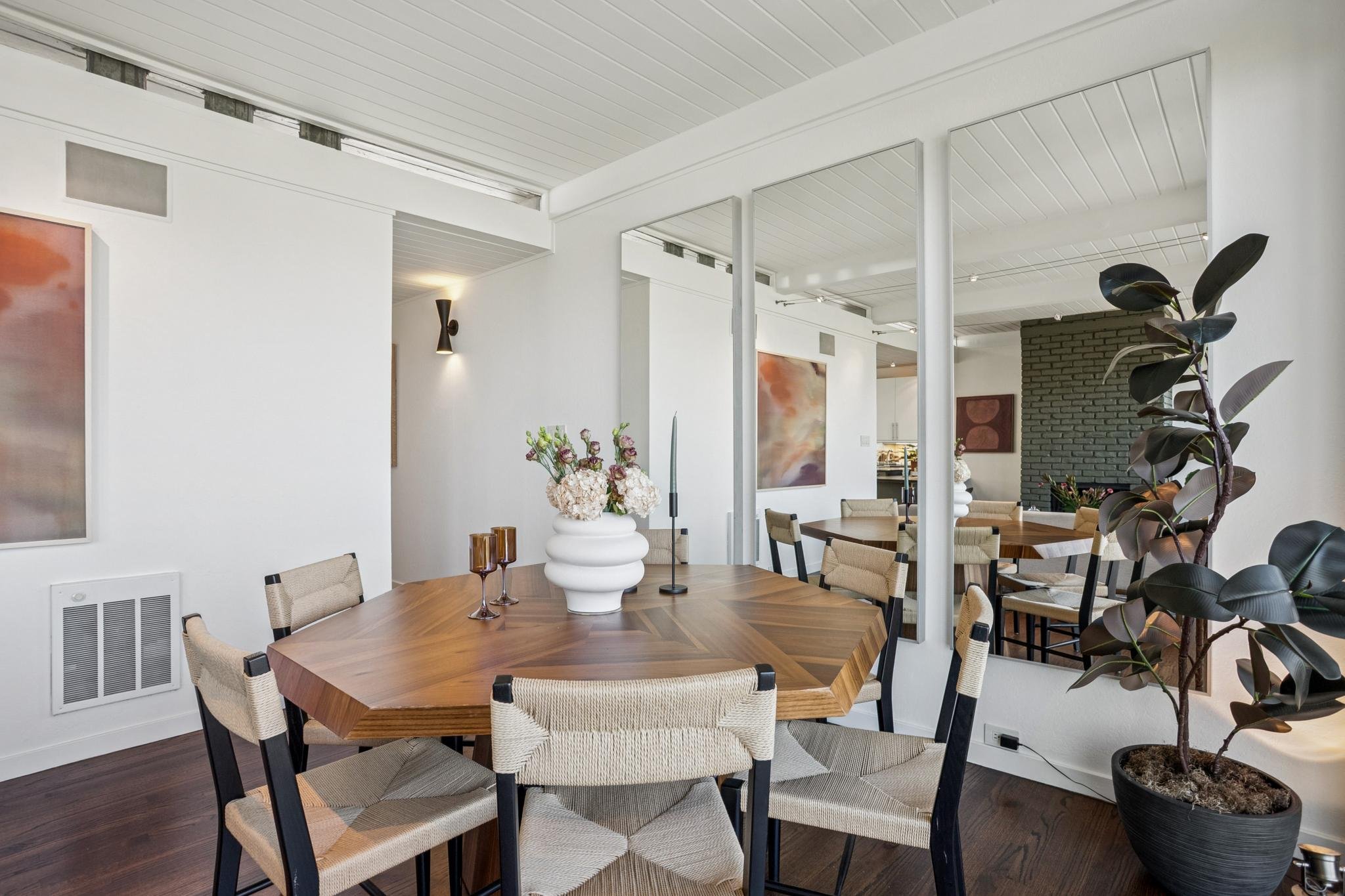 A dining room with a wooden table, beige chairs, a vase with flowers, and a large plant. The room has white walls, a white ceiling with wood paneling, and artwork on the walls.