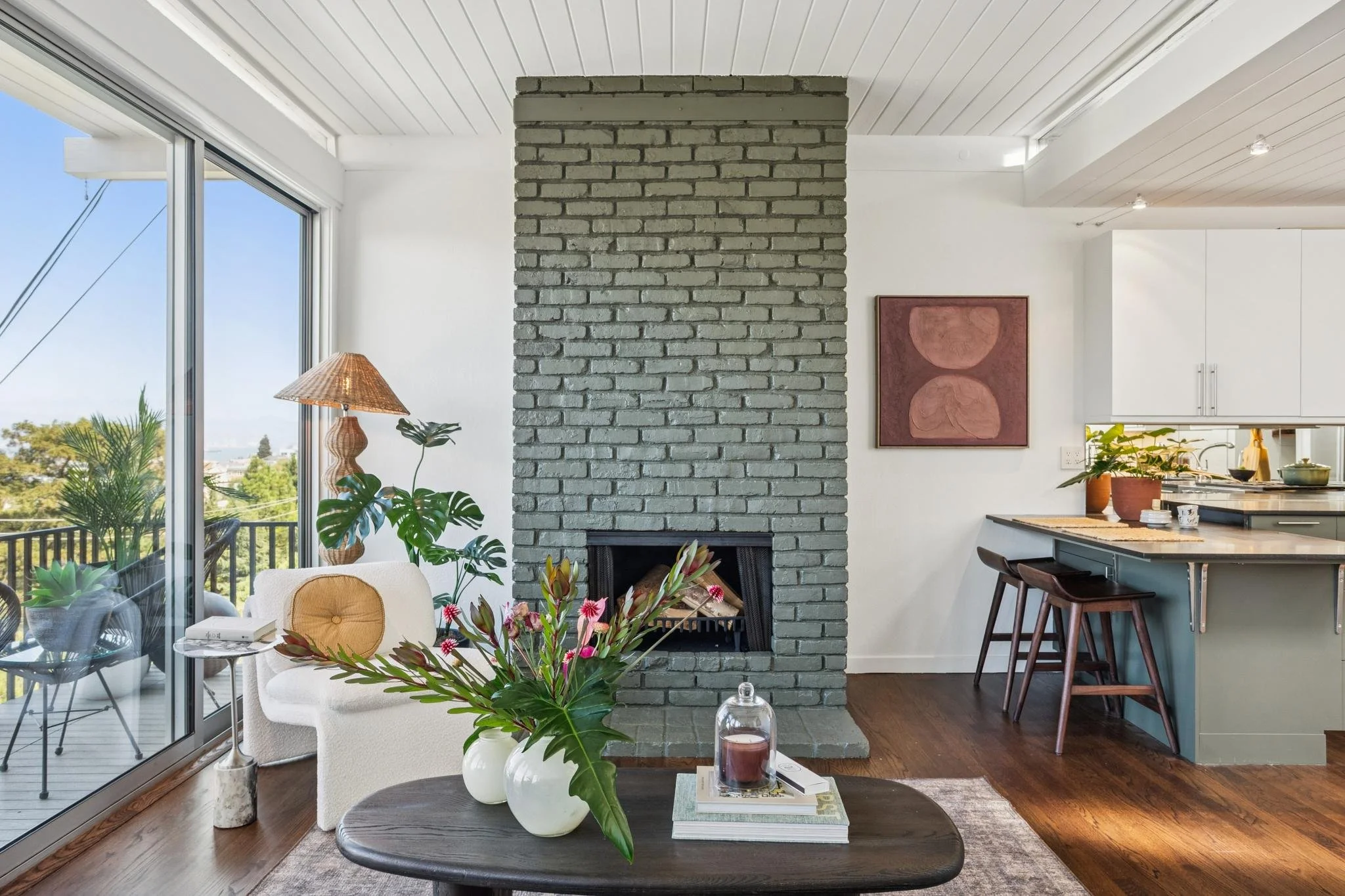 Living room with large glass sliding door, green plants, a beige chair with a mustard cushion, a black coffee table with books and a flower arrangement, a brick fireplace, and a view of a balcony with outdoor furniture and trees outside.