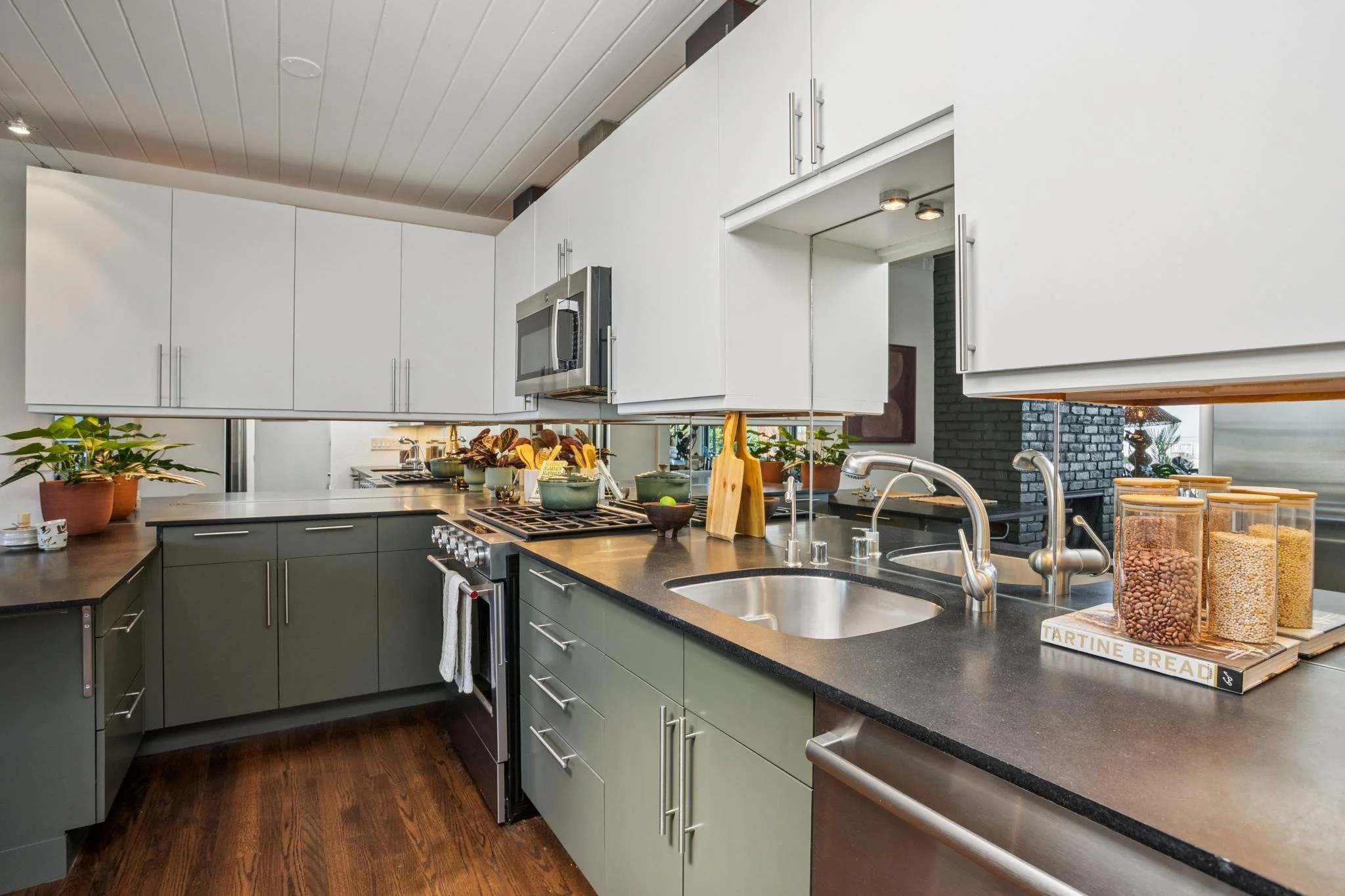 A modern kitchen with white upper cabinets, dark lower cabinets, a stainless steel microwave, and a black countertop with various kitchen items including plants, wooden spoons, and cereal jars.