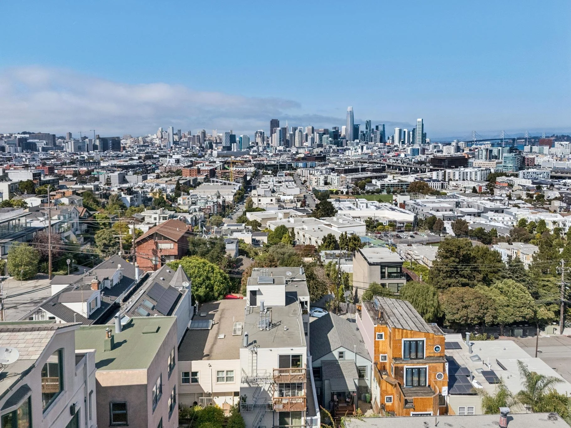 A cityscape of San Francisco, California, featuring various residential and commercial buildings in the foreground and a downtown skyline with high-rise buildings in the background, including the Salesforce Tower, with the San Francisco Bay and the B