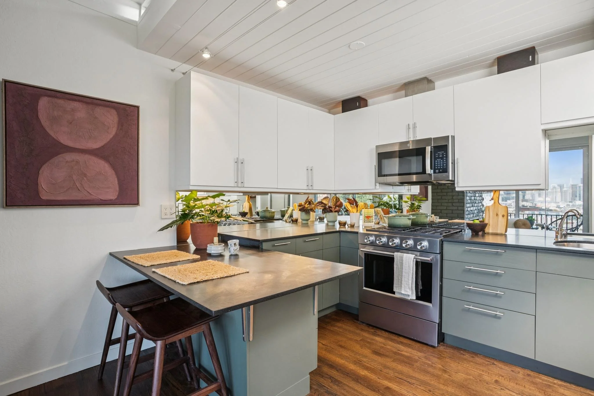Modern kitchen with white cabinets, stainless steel microwave, stove, and dark green lower cabinets, decorated with potted plants and artwork, overlooking city buildings through a window.