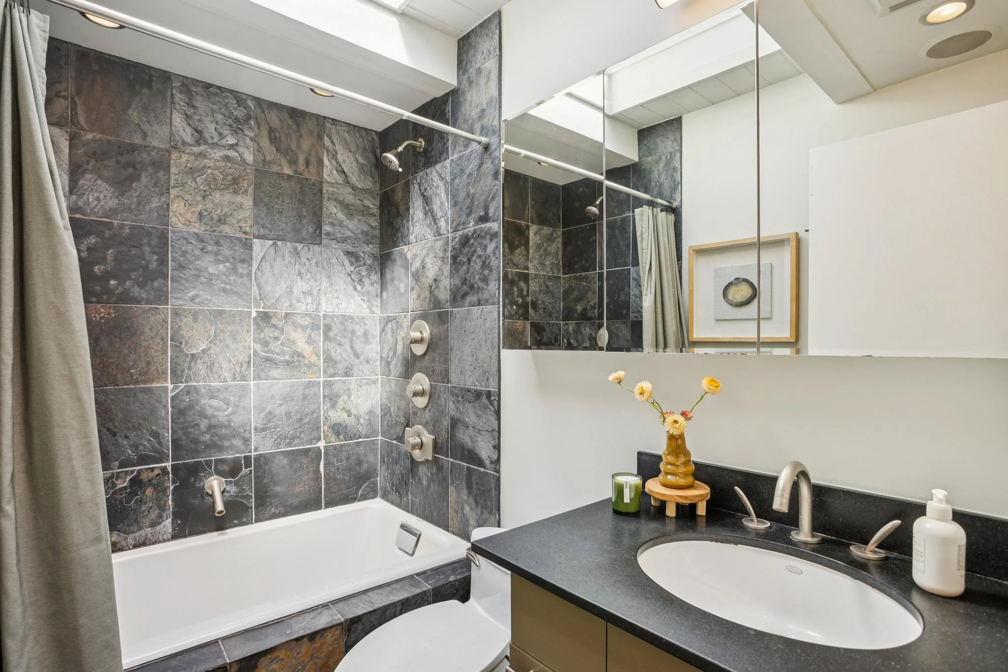 A modern bathroom with slate tile walls and a white bathtub. A shower head and controls are mounted on the wall above the bathtub. A large mirror reflects the room, showing a framed artwork and a vase with flowers on the black countertop. The bathroo