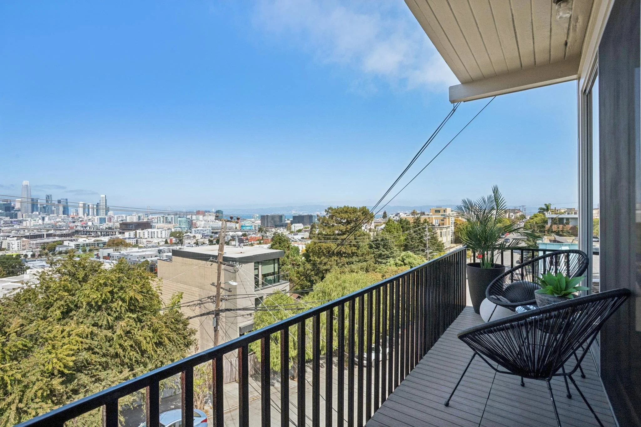 A balcony with chairs and a potted plant overlooking a cityscape with tall buildings, trees, and power lines; a blue sky with some clouds.
