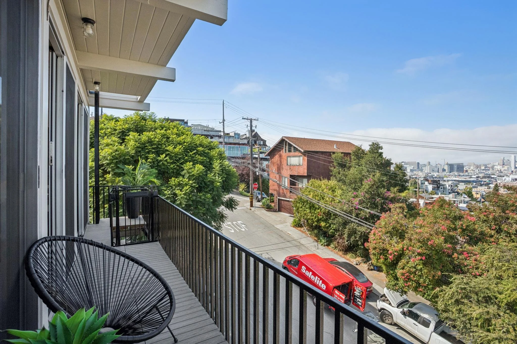 View from a balcony overlooking a city's neighborhood street with trees, parked cars, and residential buildings, with a clear blue sky above.