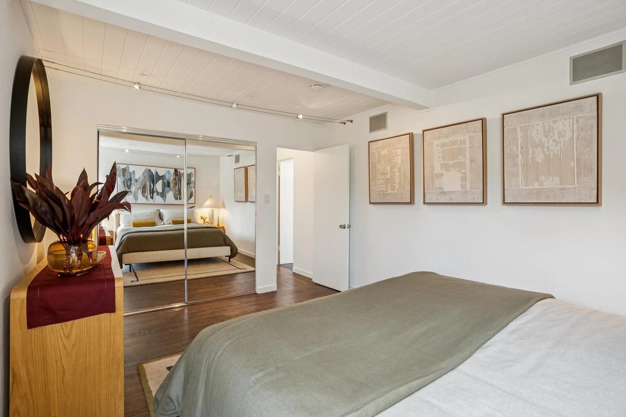 A bedroom with a bed in the foreground and an adjacent living area with a couch and artwork on the wall, separated by a mirrored closet door.