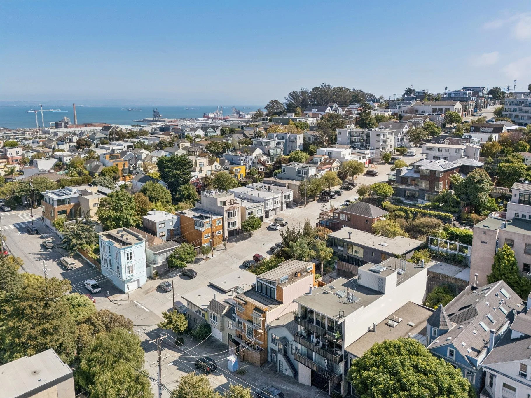 An aerial view of a coastal residential neighborhood with multicolored houses, green trees, and streets, overlooking the ocean with ships and cranes on the distant port.