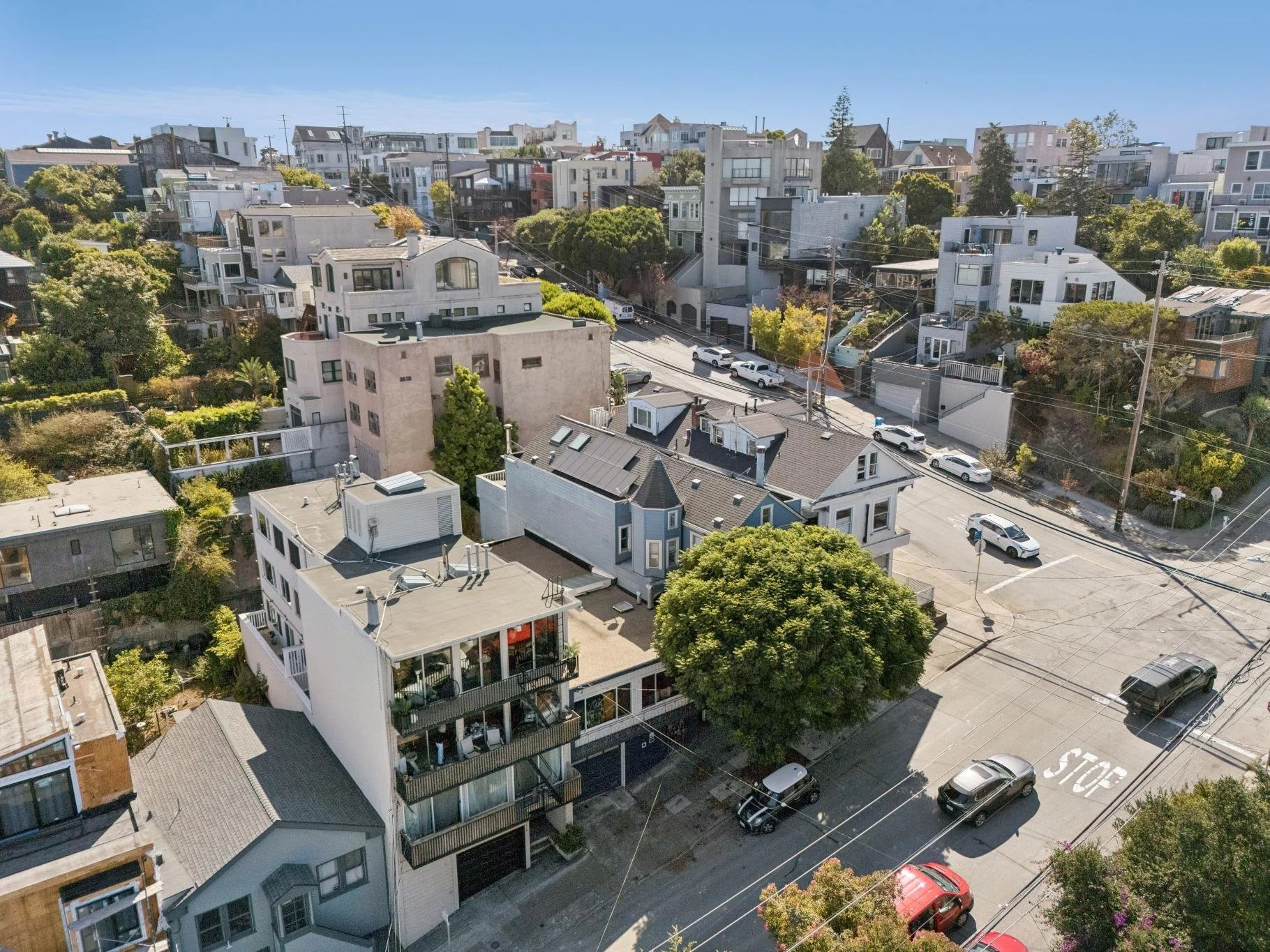 Aerial view of a neighborhood with various modern and traditional houses, trees, parked cars, and power lines on a sunny day.