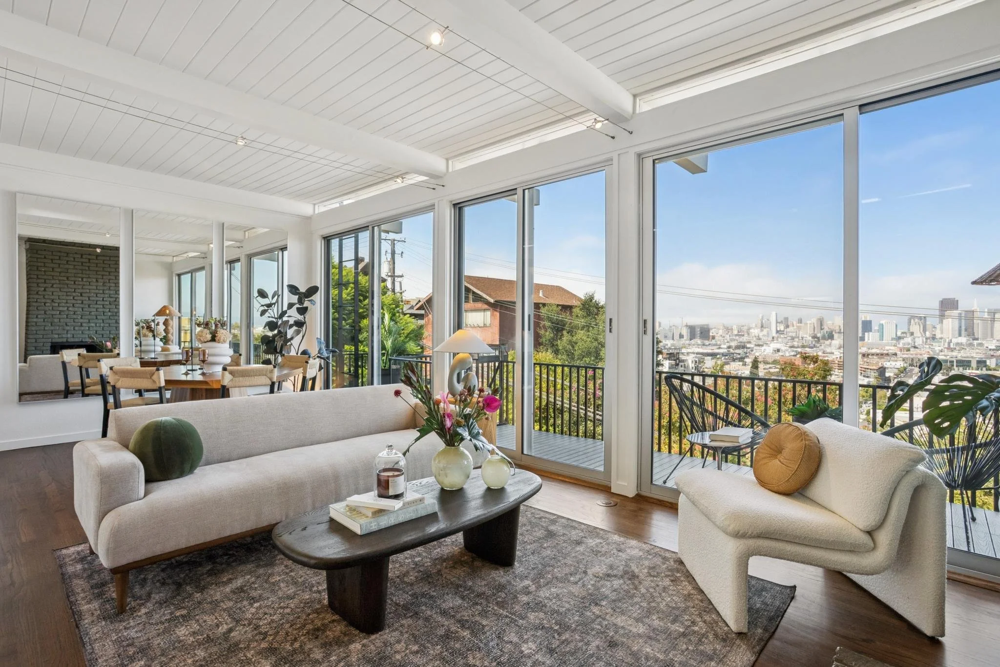 Living room with white sofas, a wooden coffee table with vases and books, large windows showing a city skyline, and a balcony with outdoor furniture.