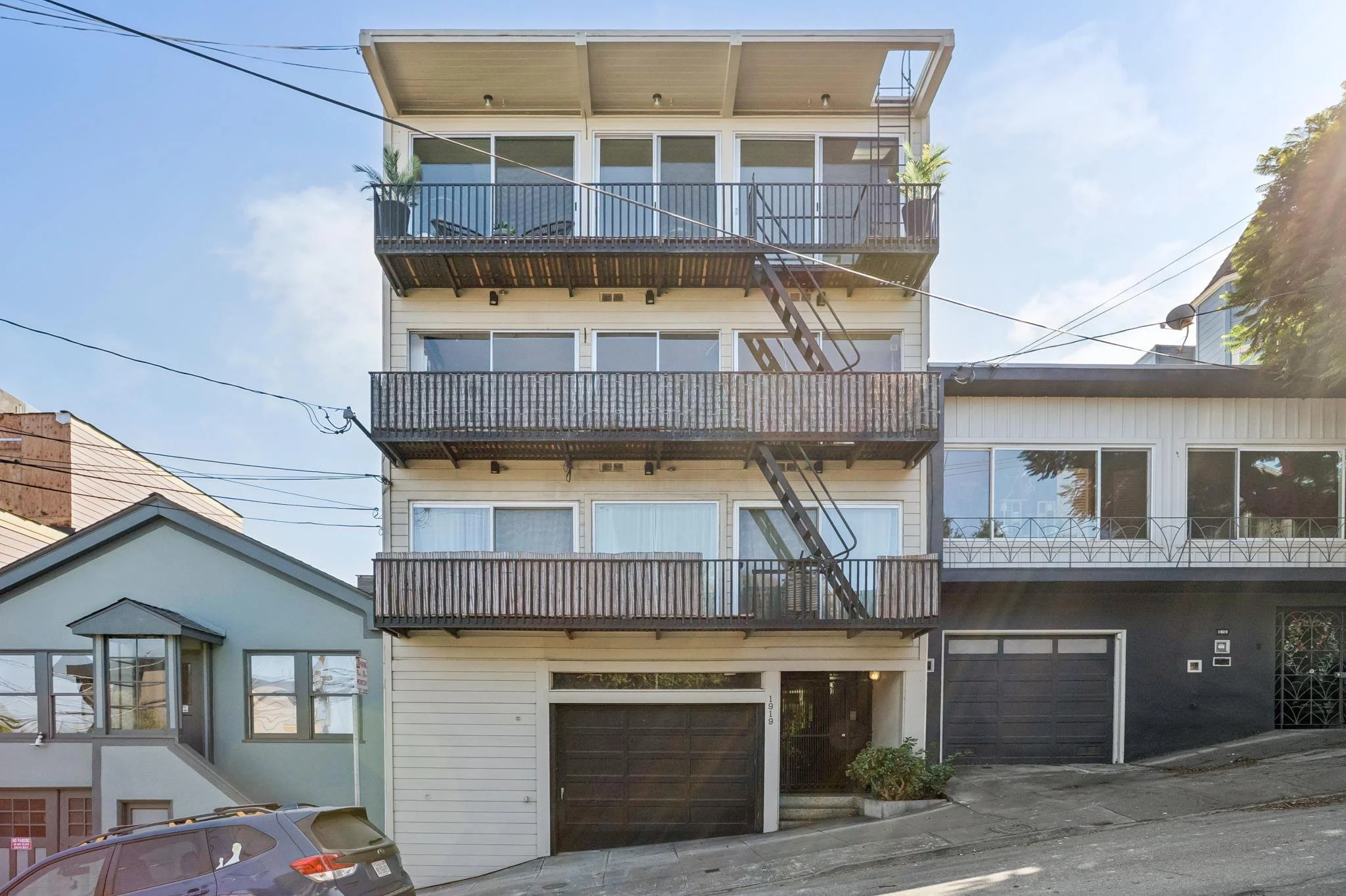 Multi-story residential building with three balconies, outdoor stairs, and a black garage door. A car is parked in front, and there are power lines overhead.