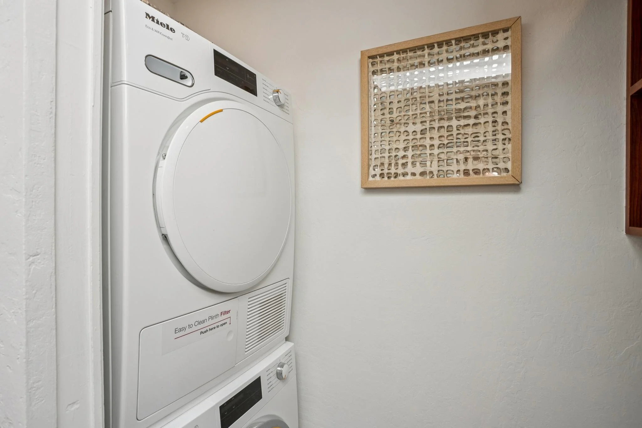 A stacked white Miele dryer and washer with control panels, placed against a plain white wall. There is a decorative wooden frame on the wall to the right of the appliances.