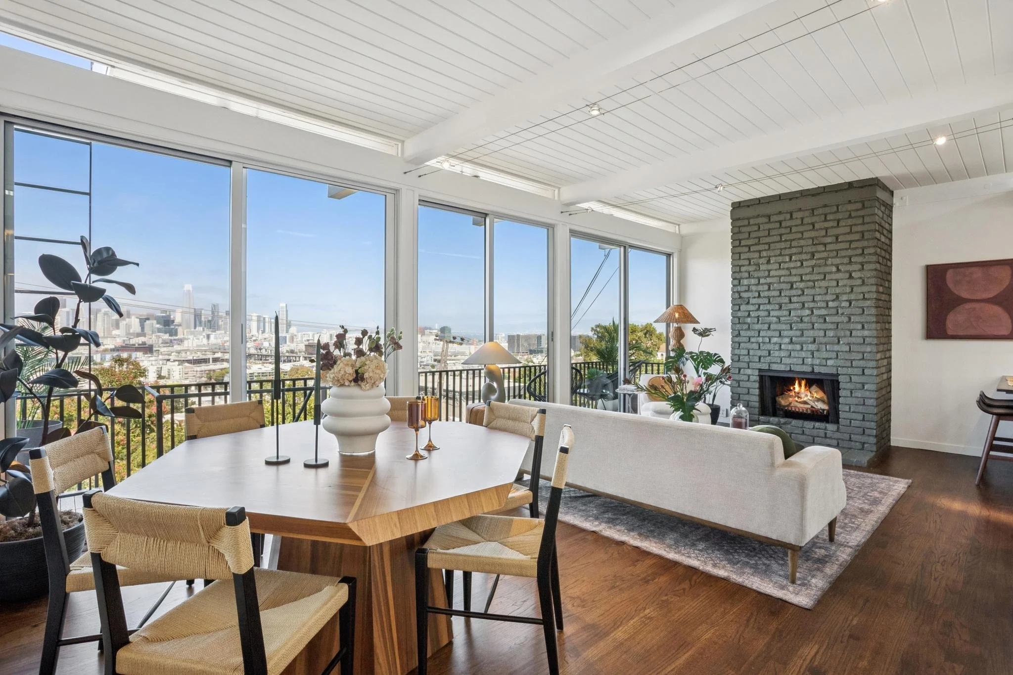 Modern living and dining room with large windows, city skyline view, brick fireplace, plants, and contemporary decor.