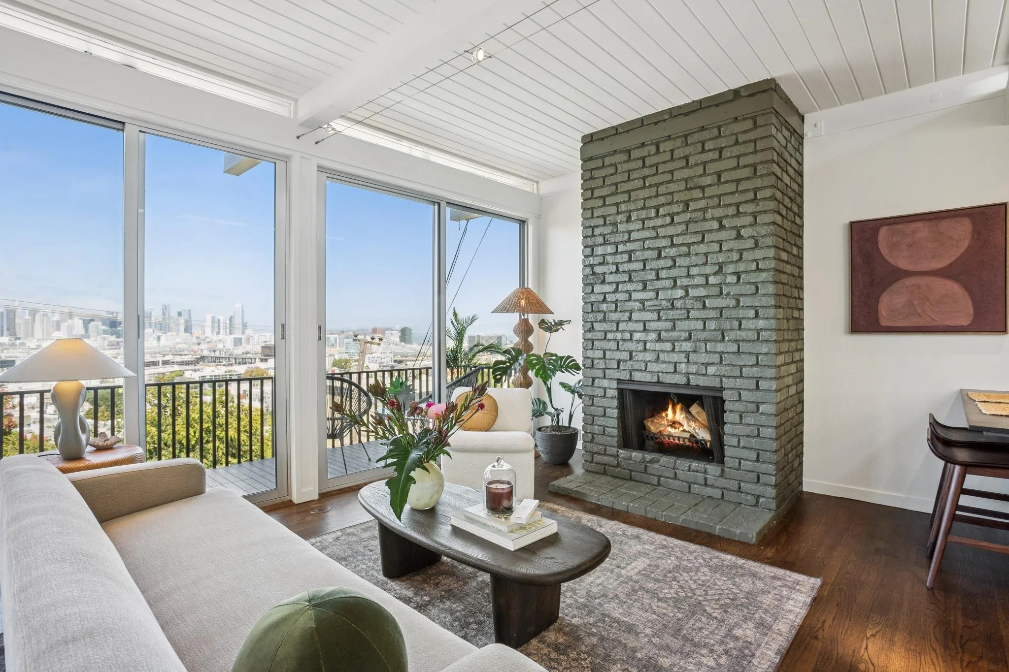 Living room with large windows showing city view, a fireplace with a chimney, a white sofa, a wooden coffee table with decorative items, plants, a side table with a lamp, and wall art.