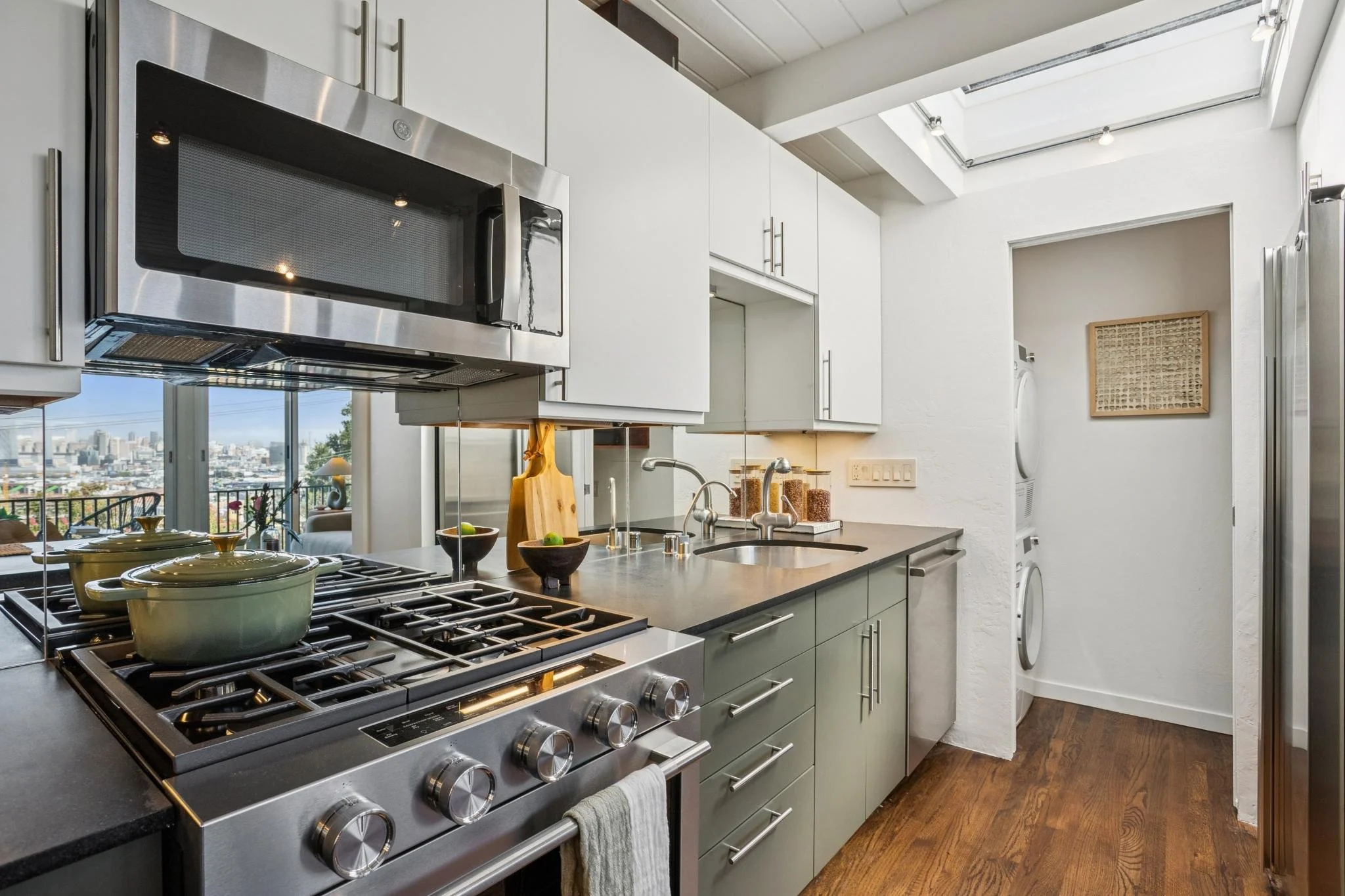 A modern kitchen with a stainless steel stove featuring multiple burners, some with pots, and an overhead microwave. There are white cabinets, a double sink, and a washer and dryer in a small adjacent laundry area. A skylight provides natural light, 