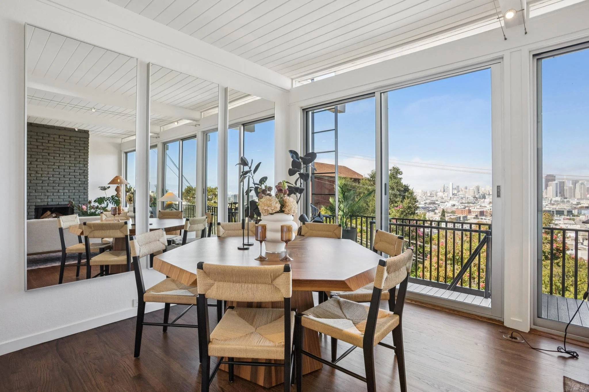 Interior of a modern dining room with a wooden table, beige chairs, a large floral centerpiece, and floor-to-ceiling windows offering city views