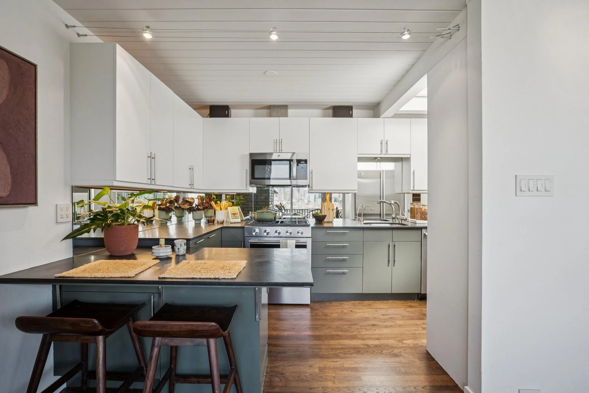 A modern kitchen with white cabinets, a stainless steel microwave, black countertops, and hardwood floors, decorated with potted plants and a couple of small rugs.