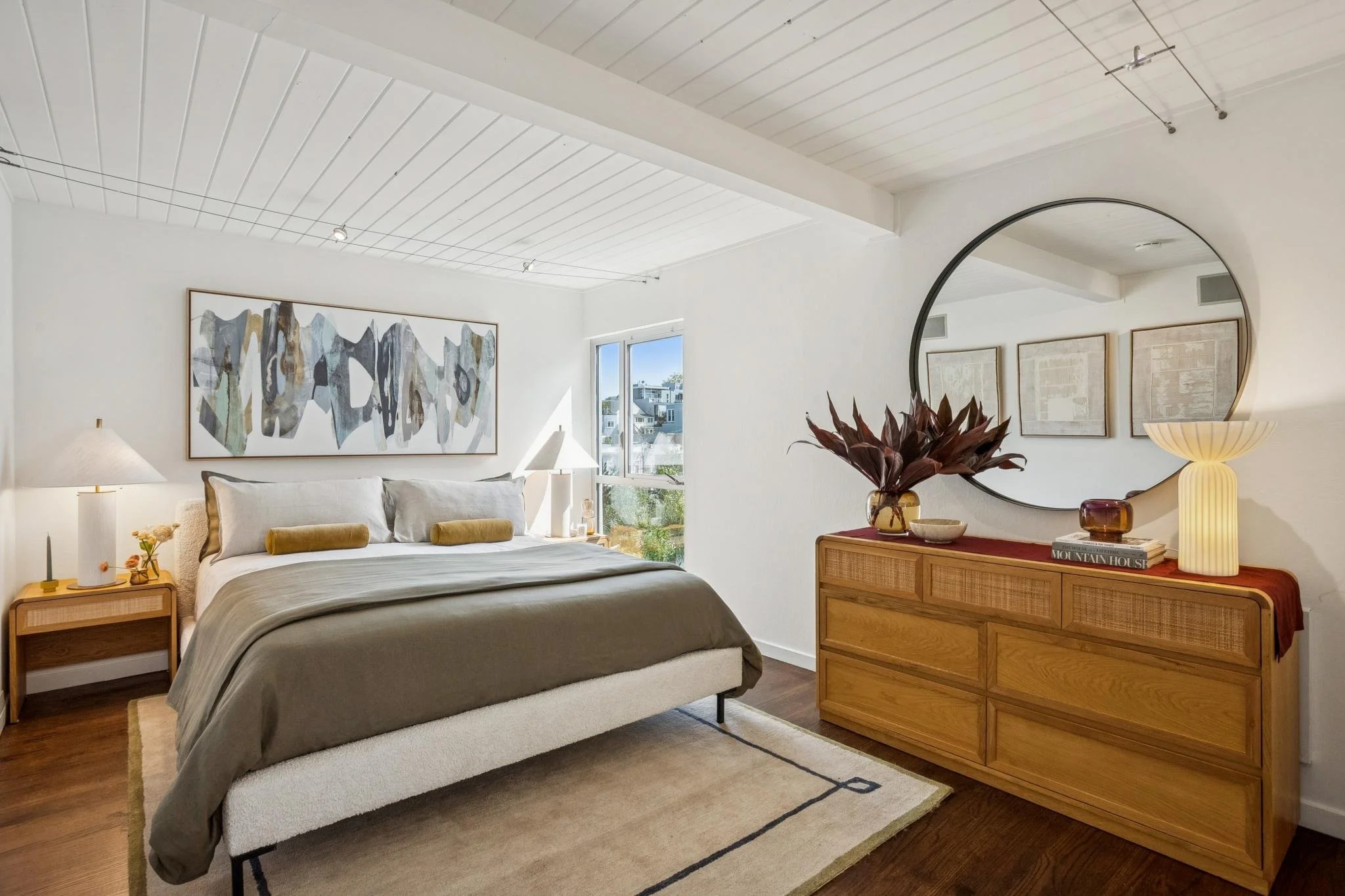 A cozy bedroom with a large bed, abstract artwork above, and natural light from a window. There are side tables with lamps and a wooden dresser with decorative items and a large round mirror.