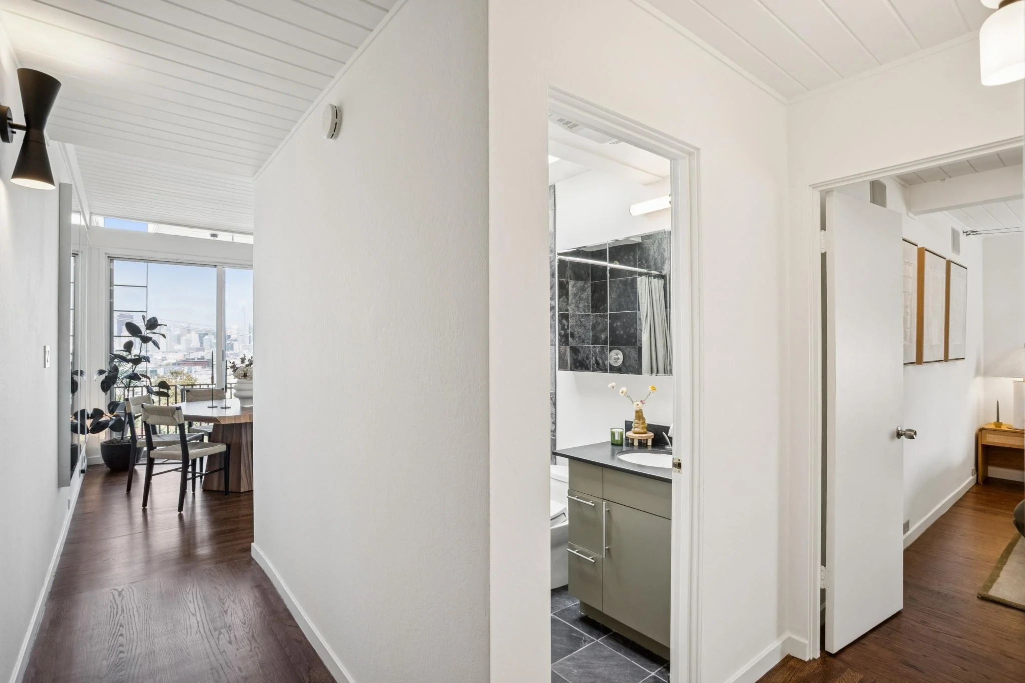 An indoor view showing a hallway with white walls and dark wooden flooring, leading to a dining area with a round table and chairs, and a nearby bathroom with dark gray tiles and a shower curtain. There's a window in the dining area offering a citysc