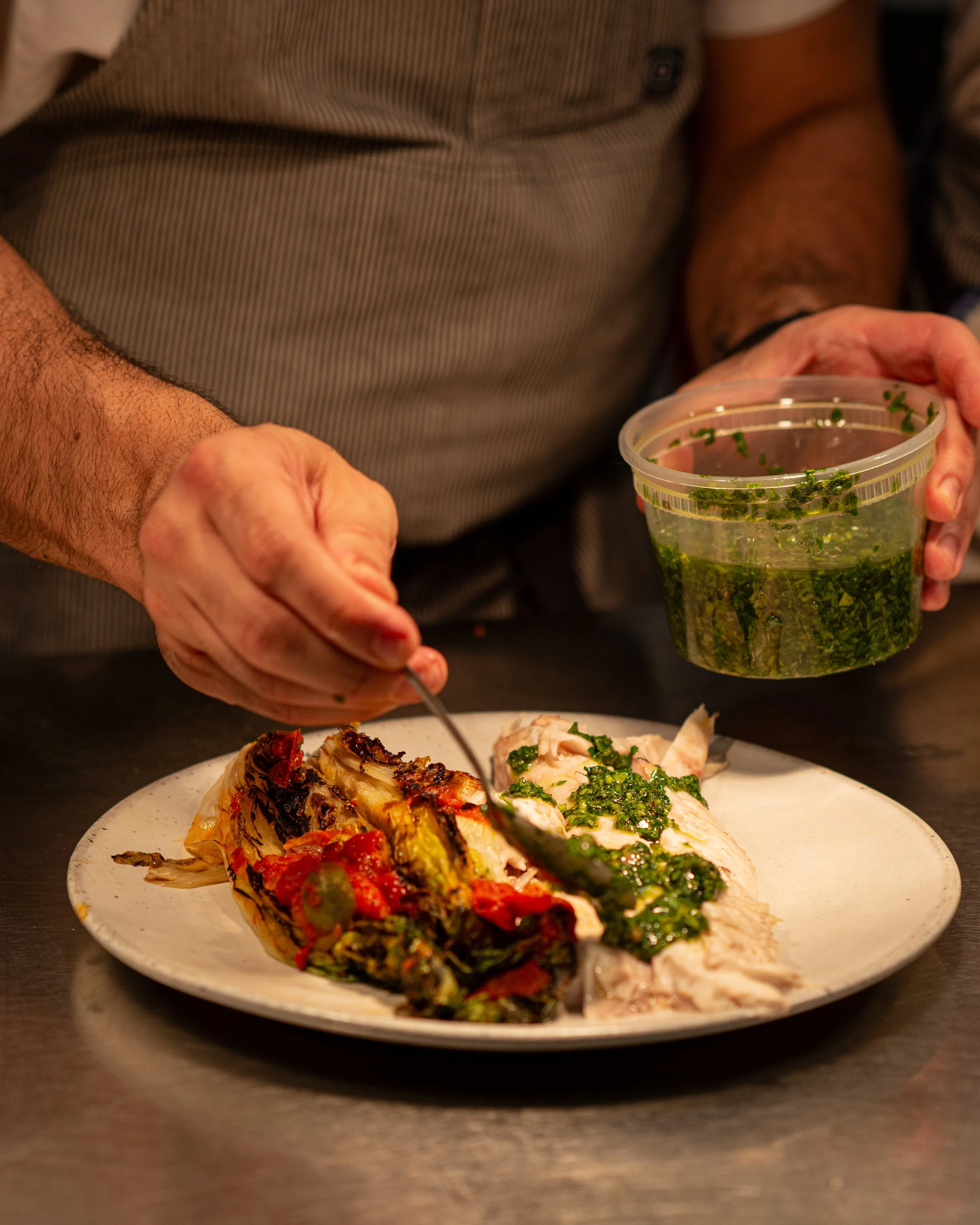 Person serving sliced grilled chicken with green herbs and roasted vegetables on a white plate. The person is holding a container with chopped herbs in one hand and a fork in the other, standing at a table.