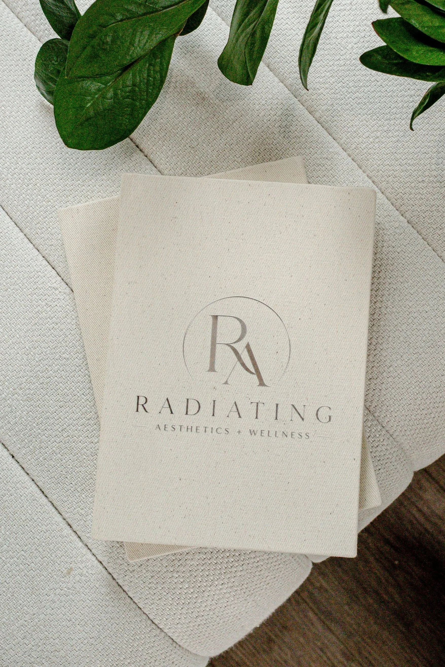 A cream-colored paper bag with a logo and text that reads "Radiance Aesthetics + Wellness" placed on a textured white surface. A green leafy plant is partially visible at the top of the image.